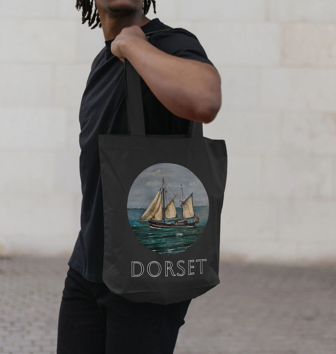 Image of a male model carrying a Black Tote Bag featuring a copy of the painting 'Moonfleet Sailing Past Portland' by Nick Dale, with the word 'Dorset' written beneath it. The bag depicts the tall ship Moonfleet sailing past the western coast of Portland, Dorset. As observed from Ringstead Beach.