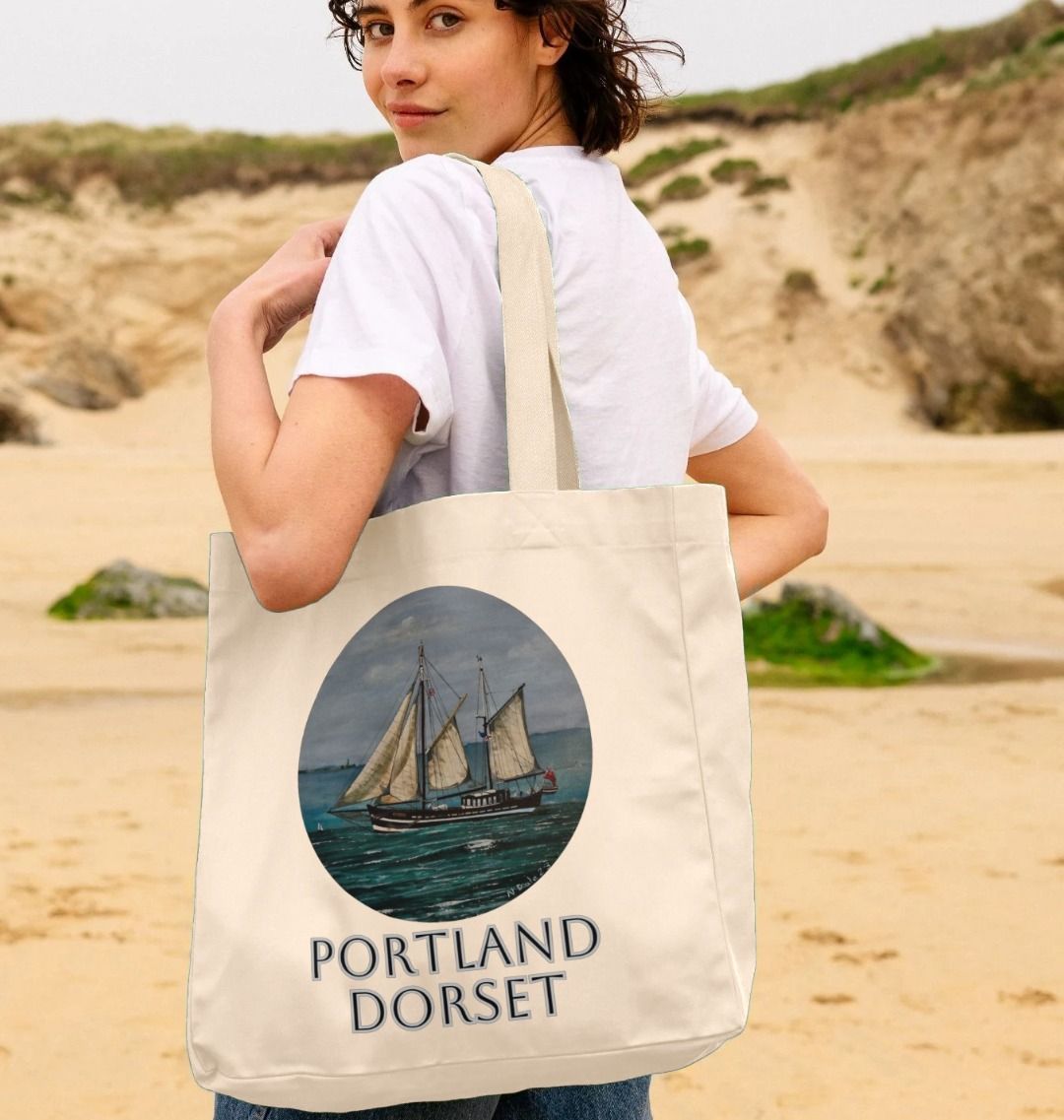 Image of a female model carrying a Natural Shopper Tote Bag featuring a copy of the painting 'Moonfleet Sailing Past Portland' by Nick Dale, with the words 'Portland Dorset' written beneath it. The bag depicts the tall ship Moonfleet sailing past the western coast of Portland, Dorset. As observed from Ringstead Beach.