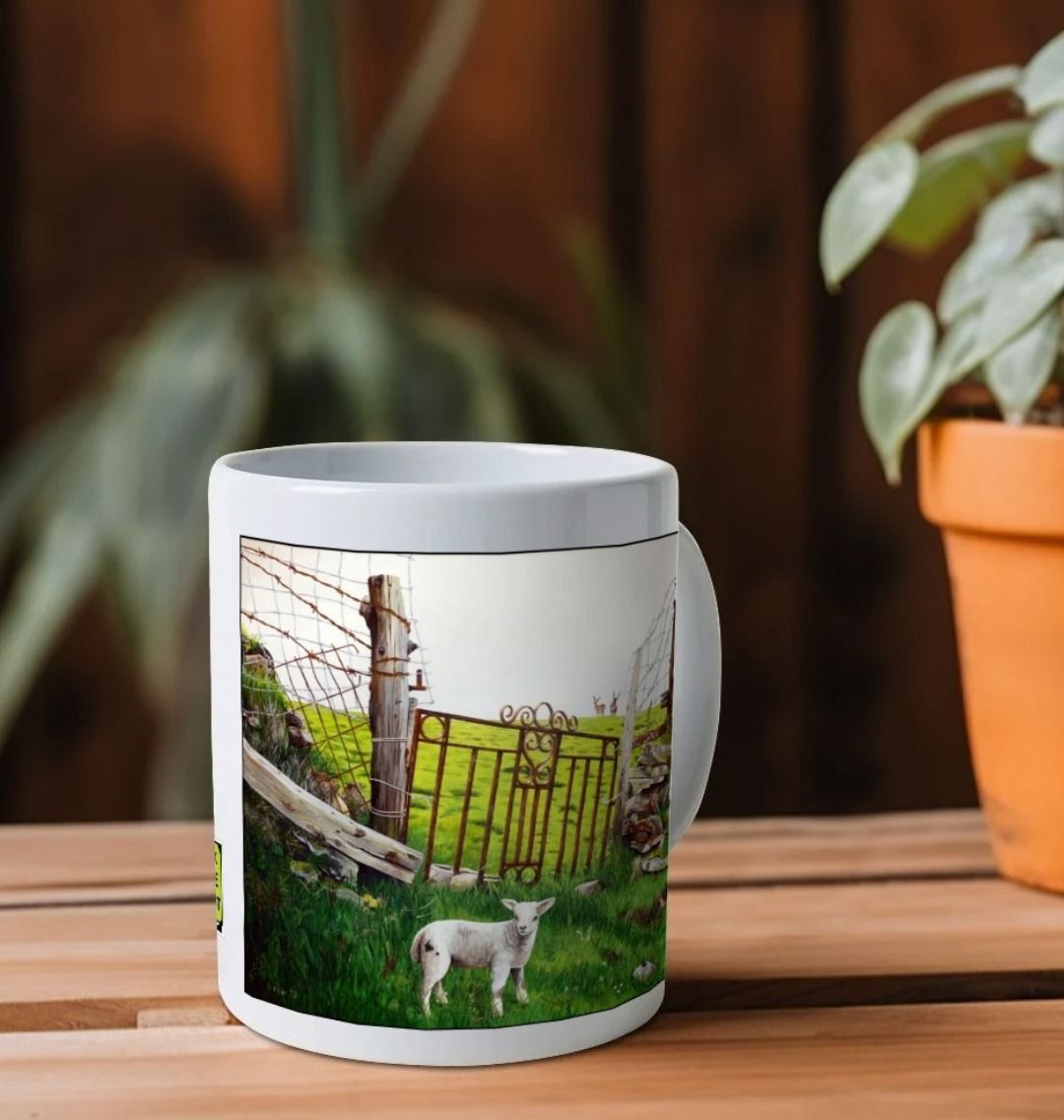 Image of a White Mug on a wooden shelf featuring a copy of the painting 'The Gate' by Nick Dale; as well as a close-up detail of a lamb from the same painting. The mug handle is obscured behind the mug. The mug depicts an ornamental garden gate reused by a farmer somewhere in North Devon, probably to the south of Valley of Rocks, Lynton. The animals depicted are all based on animals observed on Lundy Island, Devon.