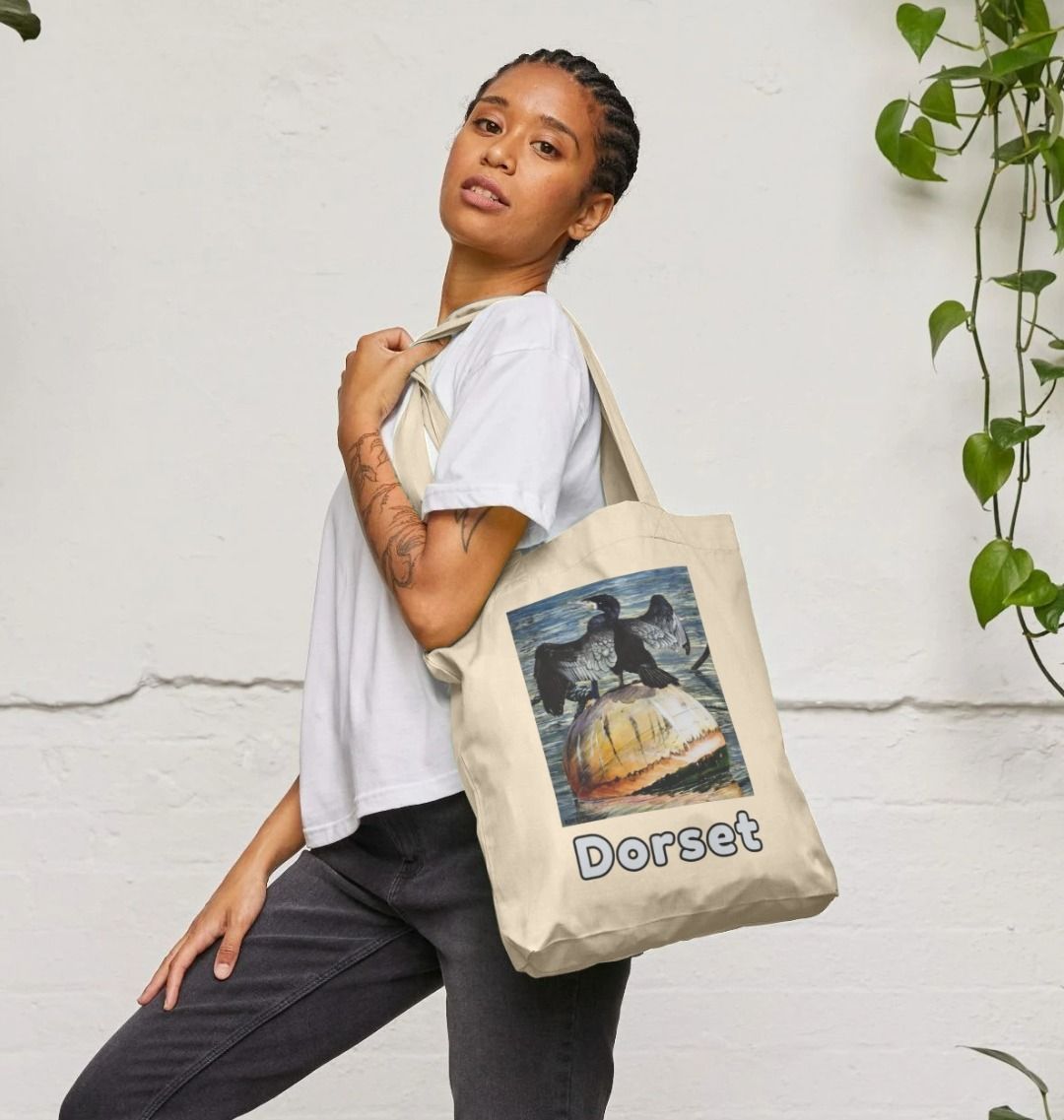 Image of a female model holding a Natural Tote Bag featuring a modified copy of the painting 'Cormorant At West Bay' by Nick Dale. The bag depicts a close-up of a cormorant sunbathing on a mooring buoy in West Bay's harbour in Dorset.