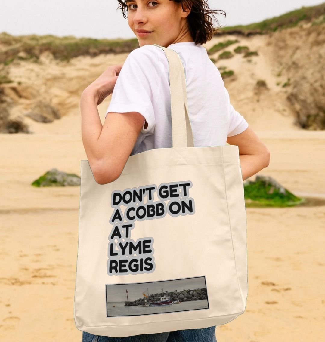 Image of a female model carrying a Natural Shopper Tote Bag featuring a copy of the painting 'End Of The Line' by Nick Dale with the slogan 'Don't Get A Cobb On At Lyme Regis' written above it. The bag depicts a fisherman preparing his catch for unloading at Lyme Regis as the birds line up on the breakwater and patiently await their turn to feed.