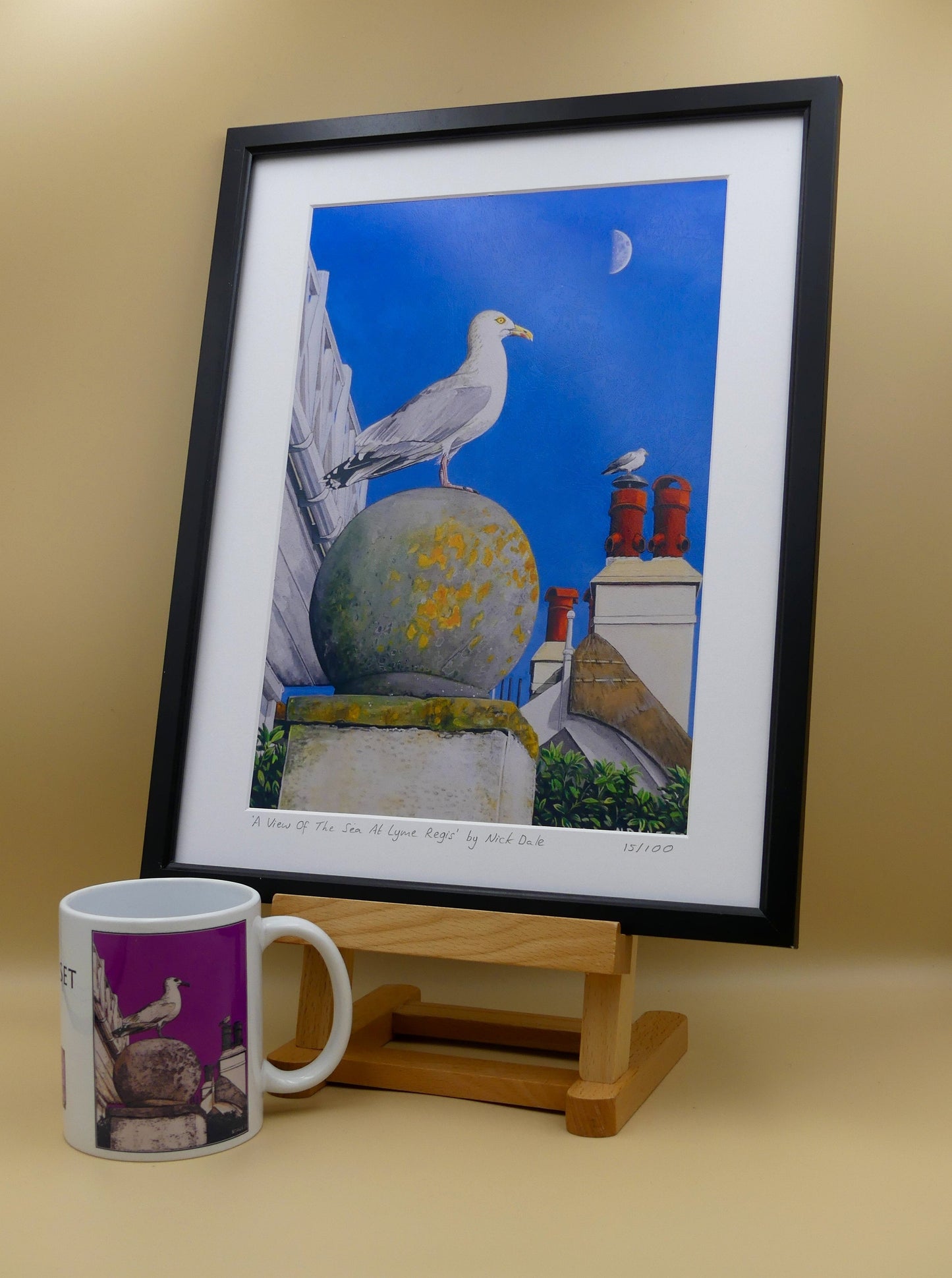 Image of Medium Art Print featuring a copy of the painting 'A View Of The Sea At Lyme Regis' by Nick Dale. The print depicts a gull and buildings along Marine Parade, Lyme Regis. The print is situated in a black frame next to a mug.
