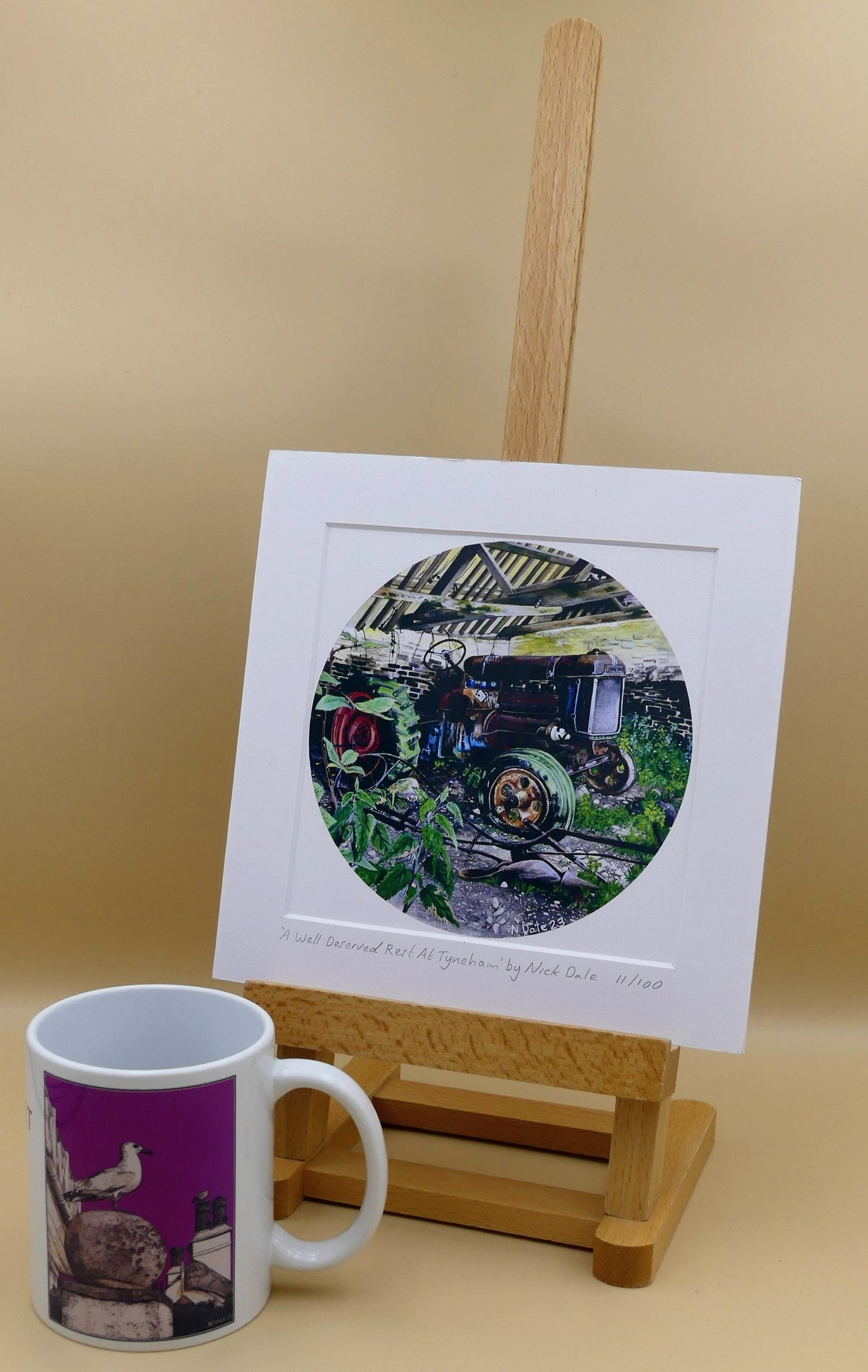 Image of Square Medium Art Print featuring a copy of the painting 'A Well Deserved Rest At Tyneham' by Nick Dale. The print depicts a Fordson Major tractor laid up in a farm building at the abandoned village of Tyneham in Dorset. The print is situated on a small easel next to a mug.