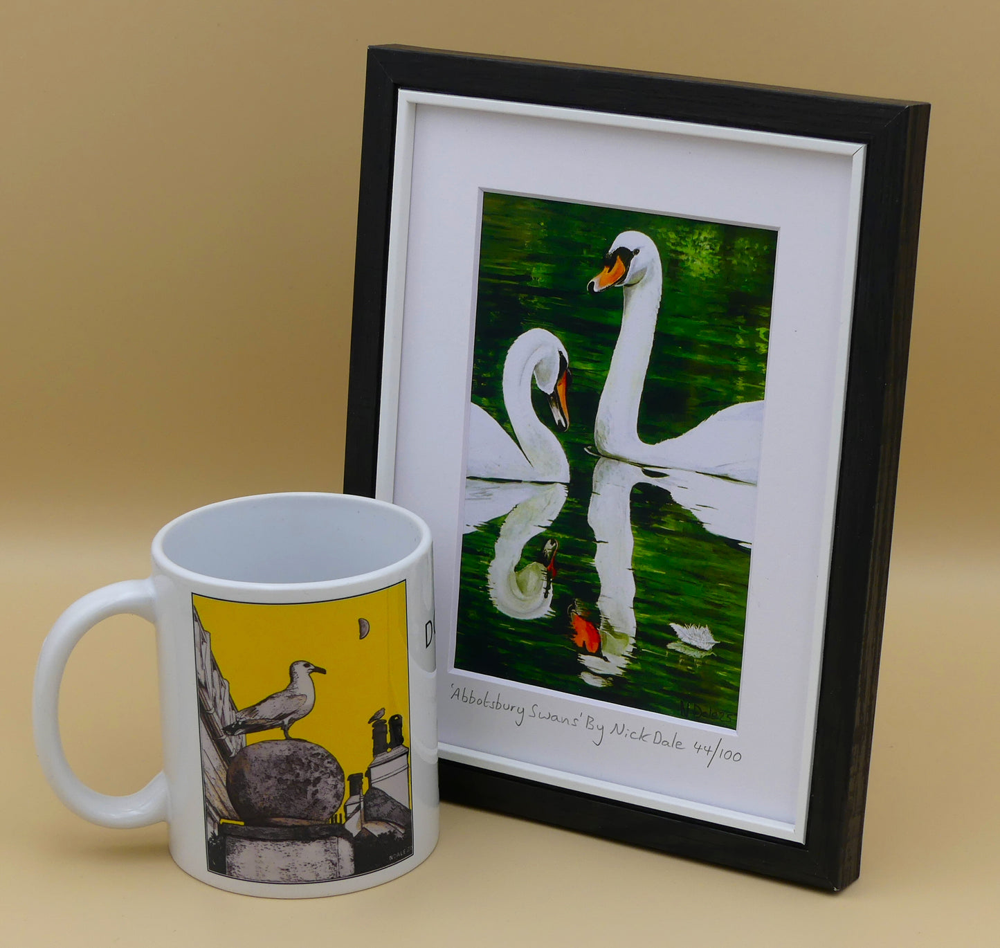 Image of a Small Art Print featuring a copy of the painting 'Abbotsbury Swans' by Nick Dale. The print depicts two swans doing the head bobbing stage of a courtship dance at Abbotsbury Swannery in Dorset. The print is situated in a black frame next to a mug.