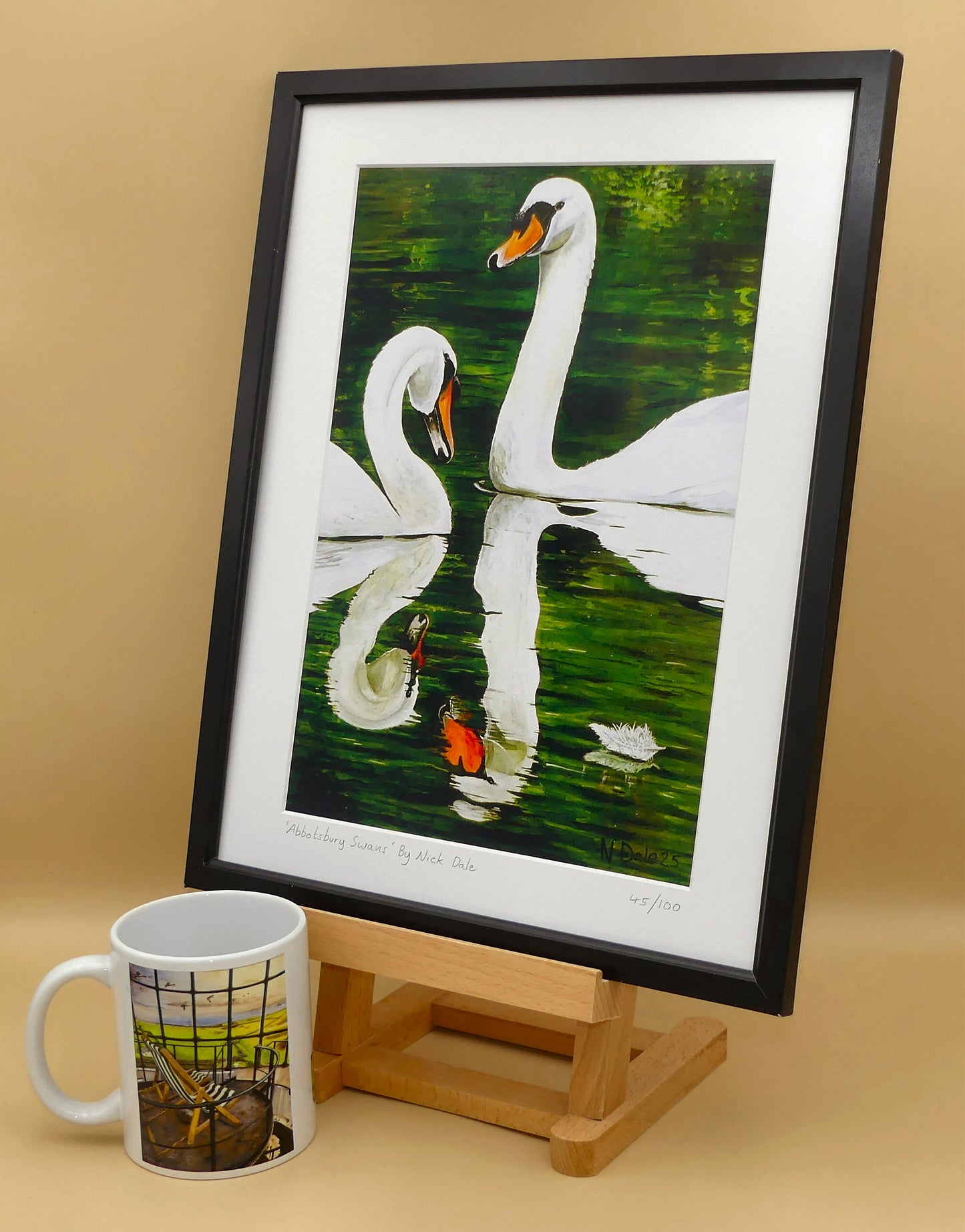 Image of a Medium Art Print featuring a copy of the painting 'Abbotsbury Swans' by Nick Dale. The print depicts two swans doing the head bobbing stage of a courtship dance at Abbotsbury Swannery in Dorset. The print is situated on a small easel in a black frame next to a mug.