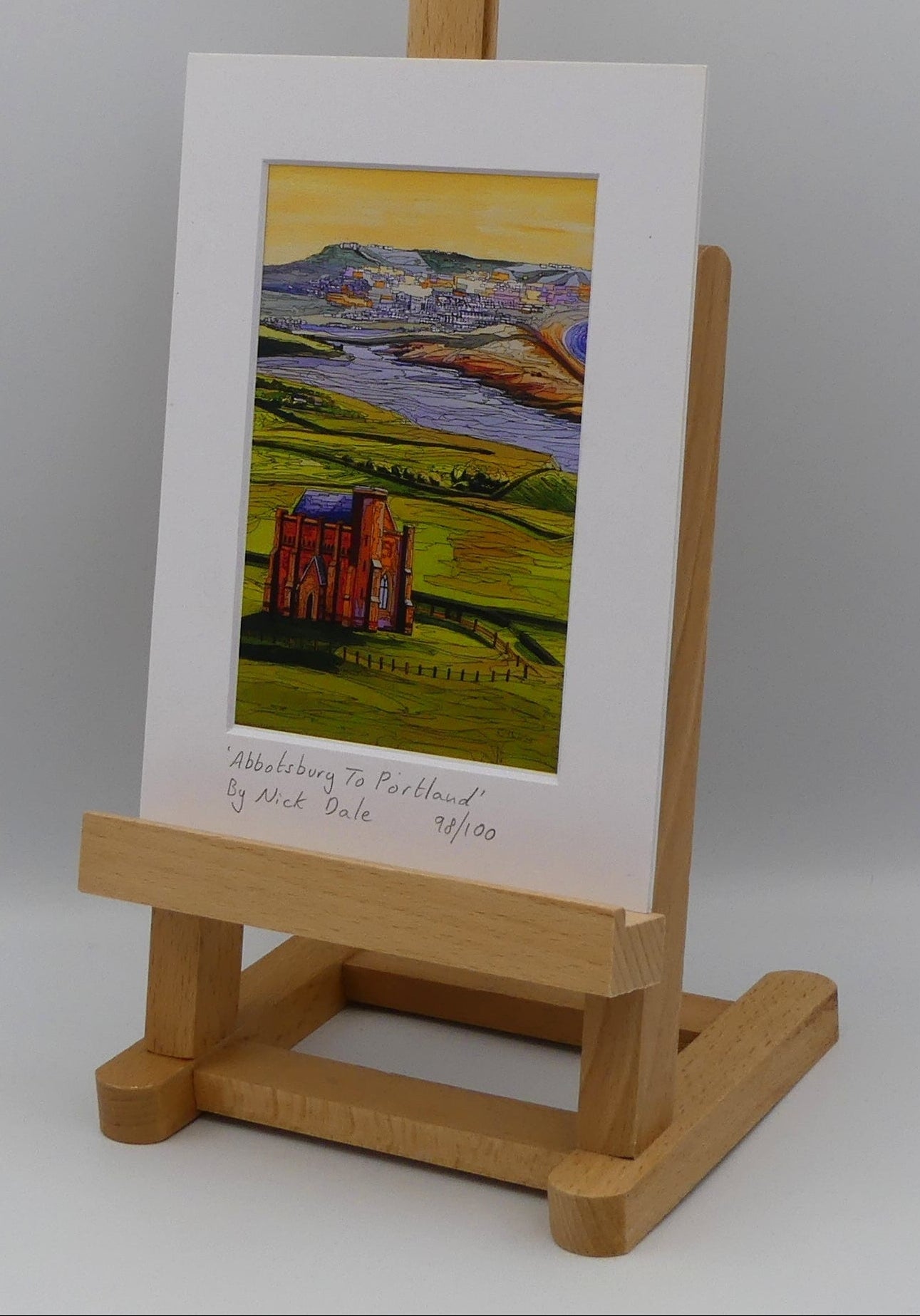 Image of a Small Art Print featuring a copy of the painting 'Abbotsbury To Portland' by Nick Dale. The print depicts the view along the Dorset coast from St Catherine's Chapel above Abbotsbury, across The Fleet lagoon to Chesil Beach and ultimately to Portland beyond it. The print is situated on a small easel.