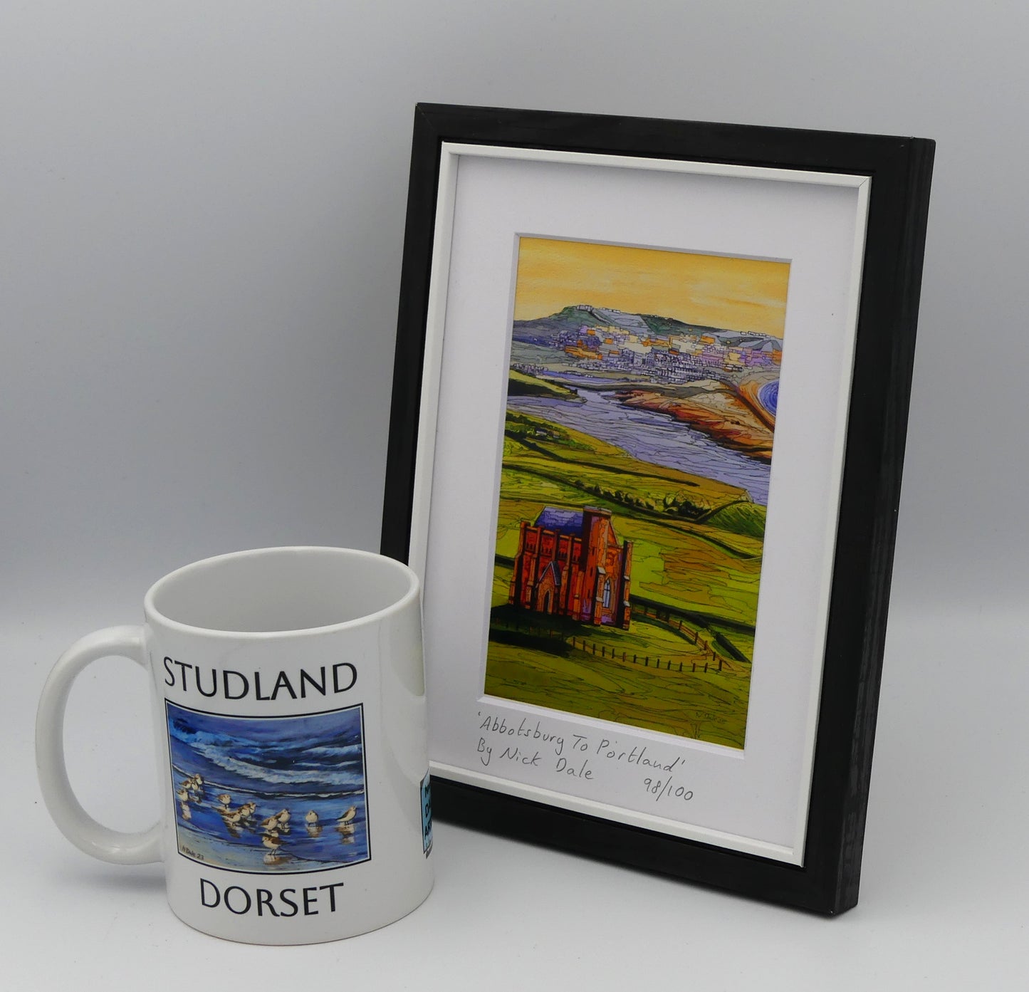 Image of a Small Art Print featuring a copy of the painting 'Abbotsbury To Portland' by Nick Dale. The print depicts the view along the Dorset coast from St Catherine's Chapel above Abbotsbury, across The Fleet lagoon to Chesil Beach and ultimately to Portland beyond it. The print is situated in a black frame next to a mug.