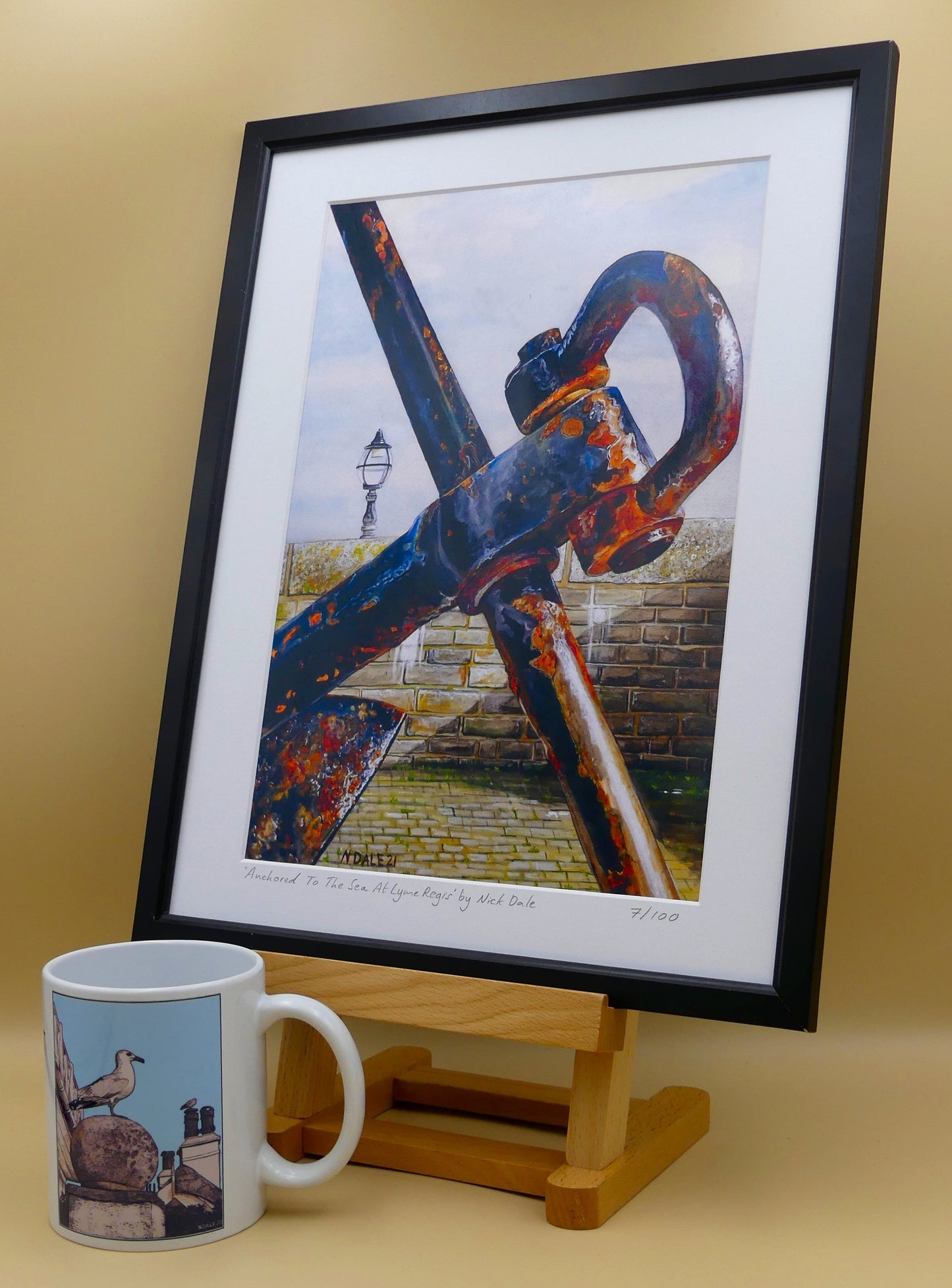 Image of Medium Art Print featuring a copy of the painting 'Anchored To The Sea At Lyme Regis' by Nick Dale. The print depicts a close up of a large anchor situated on Lyme Regis seafront along Gun Cliff Walk. The anchor was gifted from Portland Royal Naval Base in memory of all the Lyme Regis men and women who made their living from the sea. The print is situated in a black frame next to a mug.