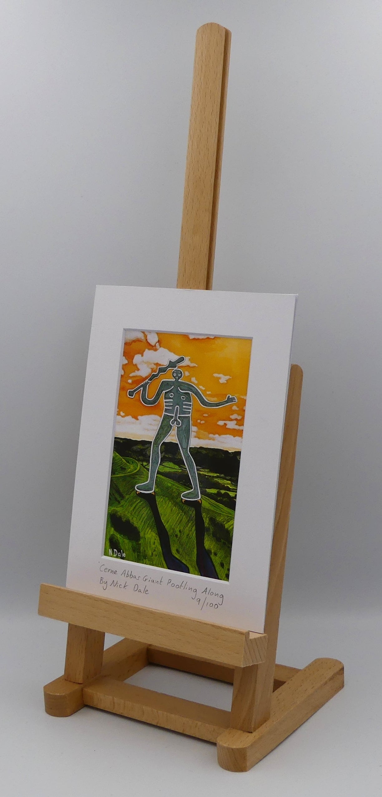 Image of a Small Art Print featuring a copy of the painting 'Cerne Abbas Giant Pootling Along' by Nick Dale. The scene imagines the Cerne Abbas Giant chalk hill figure wandering across the Dorset landscape. The giant can be seen scaling Hod Hill in the Blackmore Vale. The print is situated on a small easel.