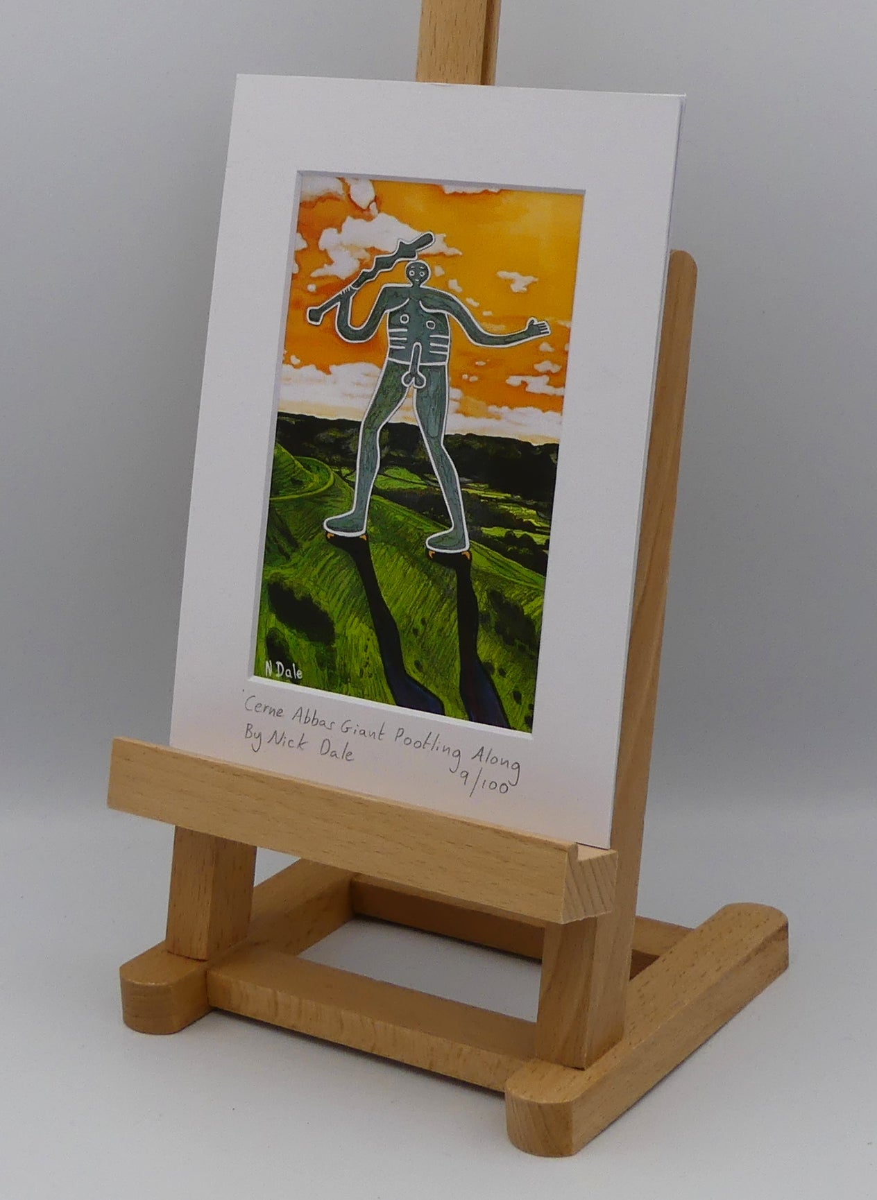 Image of a Small Art Print featuring a copy of the painting 'Cerne Abbas Giant Pootling Along' by Nick Dale. The scene imagines the Cerne Abbas Giant chalk hill figure wandering across the Dorset landscape. The giant can be seen scaling Hod Hill in the Blackmore Vale. The print is situated on a small easel.