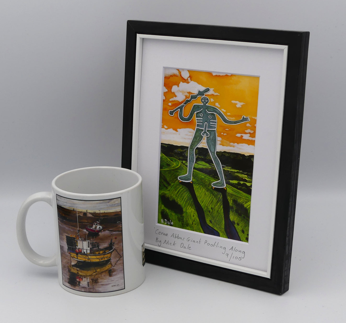 Image of a Small Art Print featuring a copy of the painting 'Cerne Abbas Giant Pootling Along' by Nick Dale. The scene imagines the Cerne Abbas Giant chalk hill figure wandering across the Dorset landscape. The giant can be seen scaling Hod Hill in the Blackmore Vale. The print is situated in a black frame next to a mug.