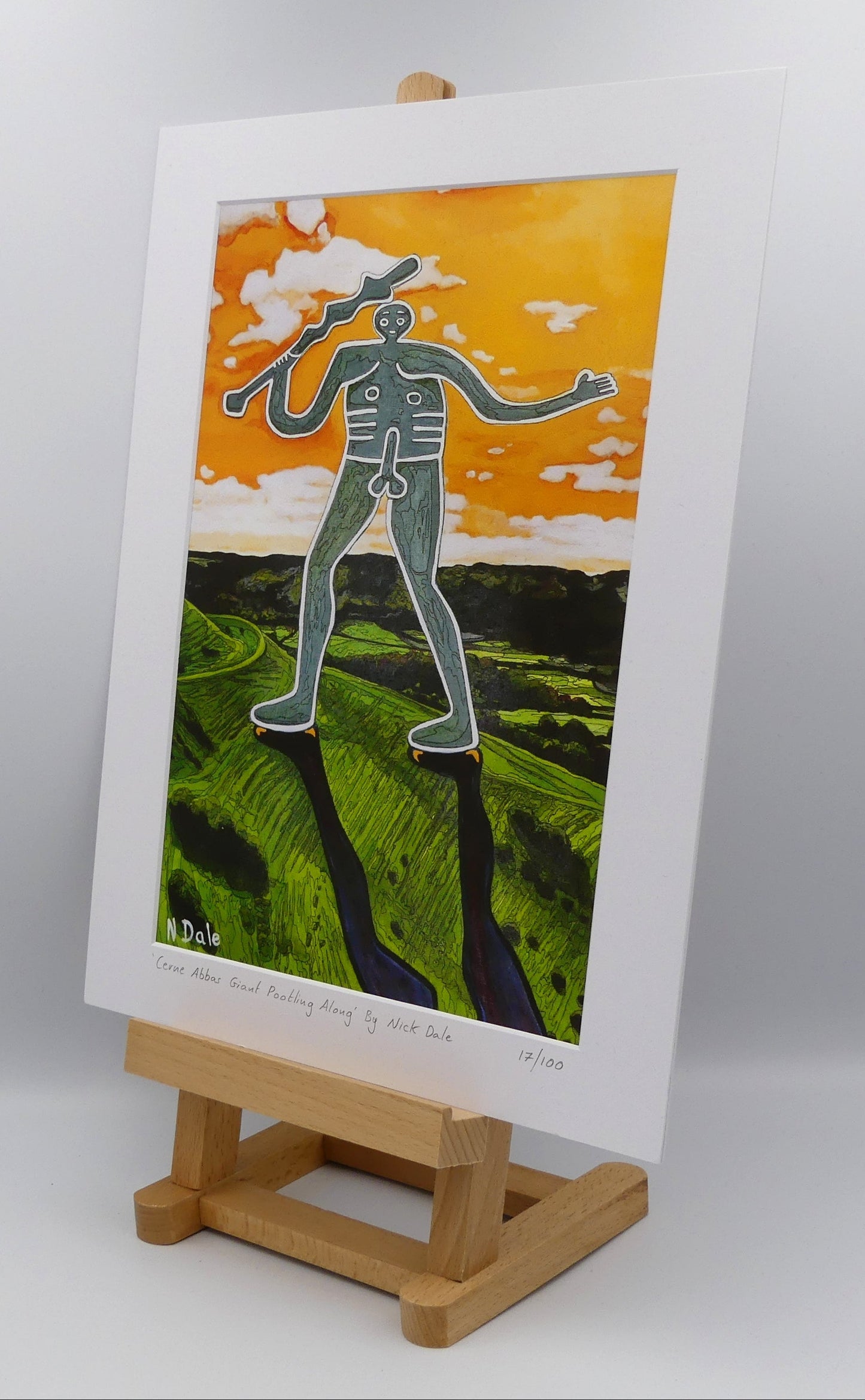 Image of a Medium Art Print featuring a copy of the painting 'Cerne Abbas Giant Pootling Along' by Nick Dale. The scene imagines the Cerne Abbas Giant chalk hill figure wandering across the Dorset landscape. The giant can be seen scaling Hod Hill in the Blackmore Vale. The print is situated on a small easel.