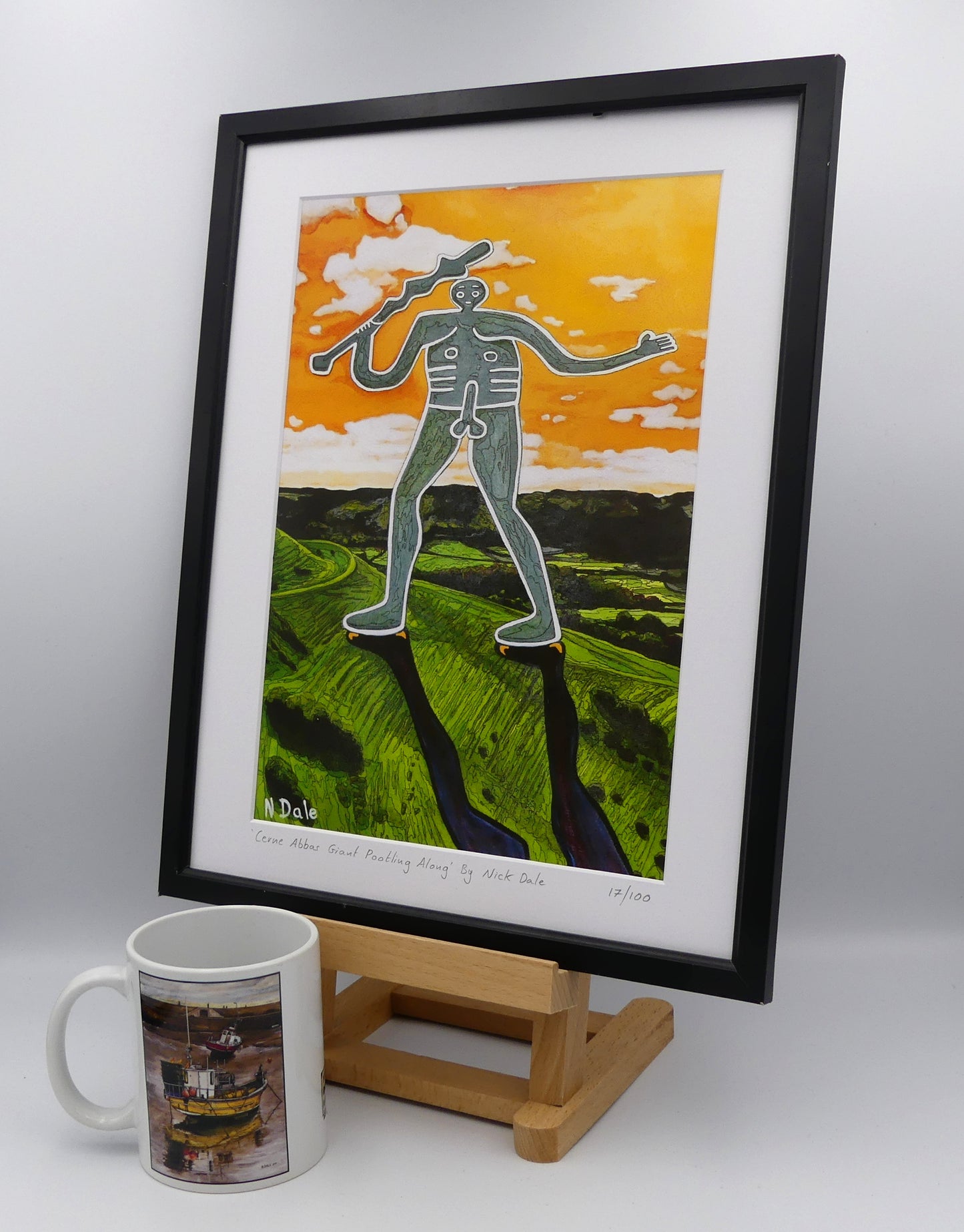 Image of a Medium Art Print featuring a copy of the painting 'Cerne Abbas Giant Pootling Along' by Nick Dale. The scene imagines the Cerne Abbas Giant chalk hill figure wandering across the Dorset landscape. The giant can be seen scaling Hod Hill in the Blackmore Vale. The print is situated on a small easel in a black frame next to a mug.