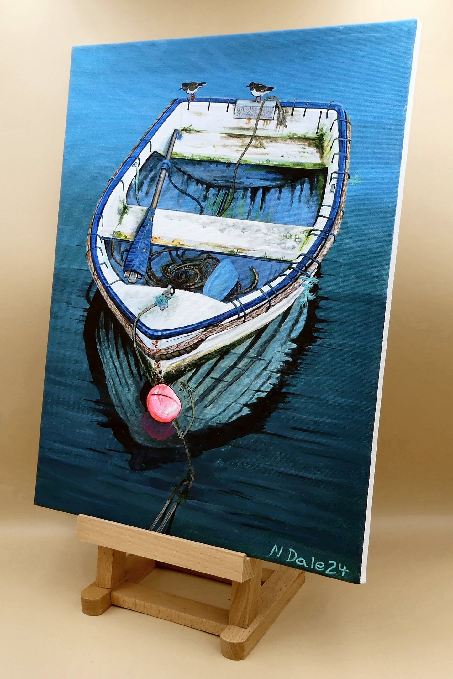Image of the original painting 'Chase Me' by Nick Dale on an easel. The painting depicts a small, somewhat neglected, rowing boat in the harbour at Lyme Regis. The title is in reference to the 2 small birds chasing each other around the edge of the boat.