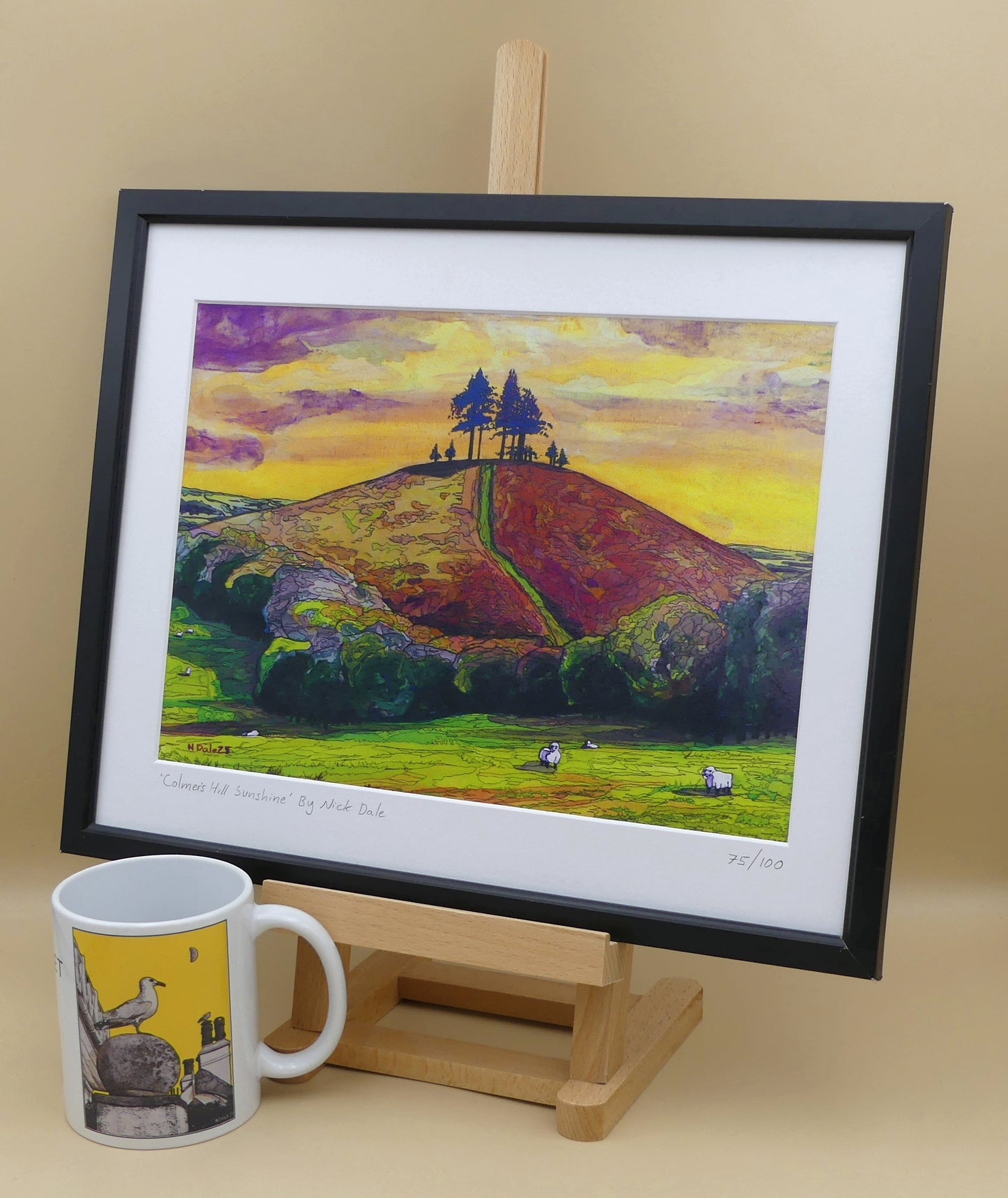 Image of a Medium Art Print featuring a copy of the painting 'Colmer's Hill Sunshine' by Nick Dale. The print depicts the prominent sandstone conical hill near the village of Symondsbury in West Dorset. The hill is topped with pine trees to commemorate those who died in World War I. The print is situated on a small easel in a black frame next to a mug.