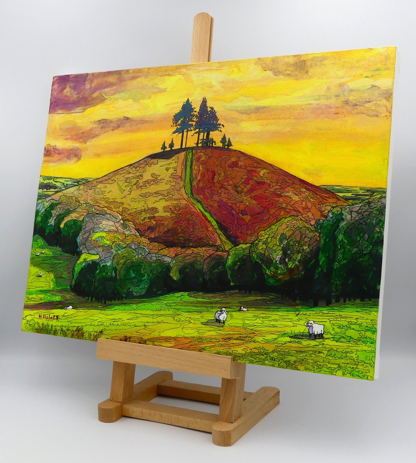 Image of the original painting 'Colmer's Hill Sunshine' by Nick Dale on an easel. The painting depicts the prominent sandstone conical hill near the village of Symondsbury in West Dorset. The hill is topped with pine trees to commemorate those who died in World War I.