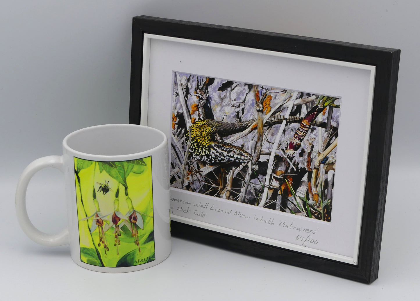 Image of a Small Art Print featuring a copy of the painting 'Common Wall Lizard Near Worth Matravers' by Nick Dale. The print depicts a close up view of a common wall lizard in the clifftop Hedbury Quarry, south of Worth Matravers. Has the lizard clocked the nearby beetle? The print is situated in a black frame next to a mug.