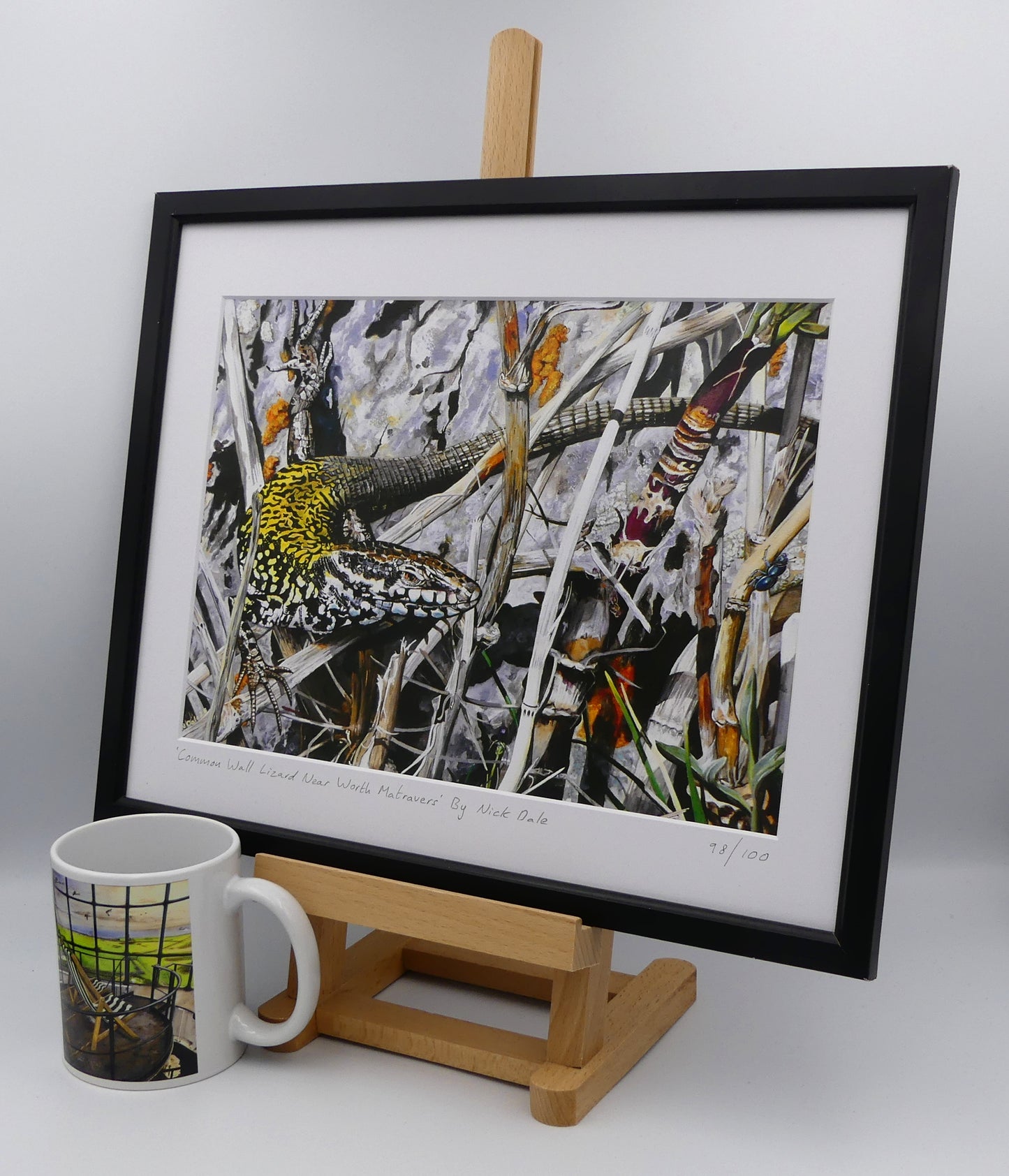 Image of a Medium Art Print featuring a copy of the painting 'Common Wall Lizard Near Worth Matravers' by Nick Dale. The print depicts a close up view of a common wall lizard in the clifftop Hedbury Quarry, south of Worth Matravers. Has the lizard clocked the nearby beetle? The print is situated on a small easel in a black frame next to a mug.