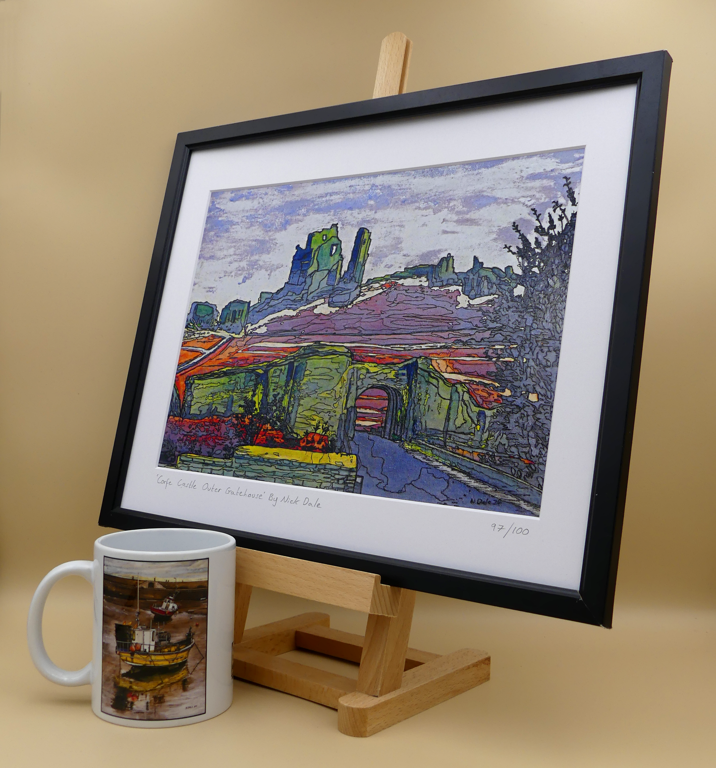 Image of a Medium Art Print featuring a copy of the painting 'Corfe Castle Outer Gatehouse' by Nick Dale. The print depicts the slighted outer gatehouse of Corfe Castle in the village of Corfe Castle, Dorset. Beyond it can be seen the castle's south-west gatehouse and keep on top of the central hilltop. The print is situated on a small easel in a black frame next to a mug.