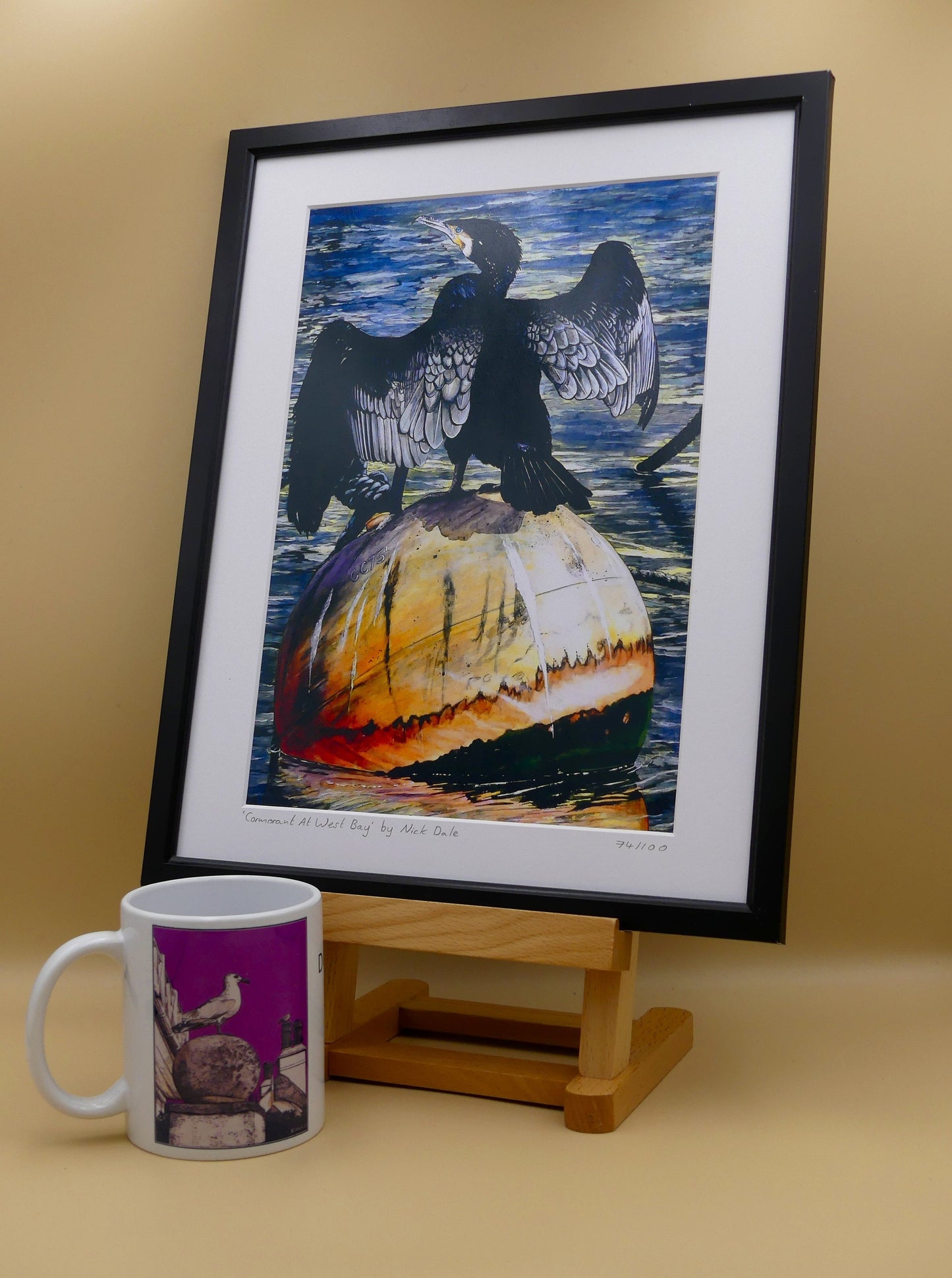 Image of Medium Art Print featuring a copy of the painting 'Cormorant At West Bay' by Nick Dale. The print depicts a close-up of a cormorant sunbathing on a Mooring Buoy in West Bay's harbour in Dorset. The print is situated in a black frame next to a mug.