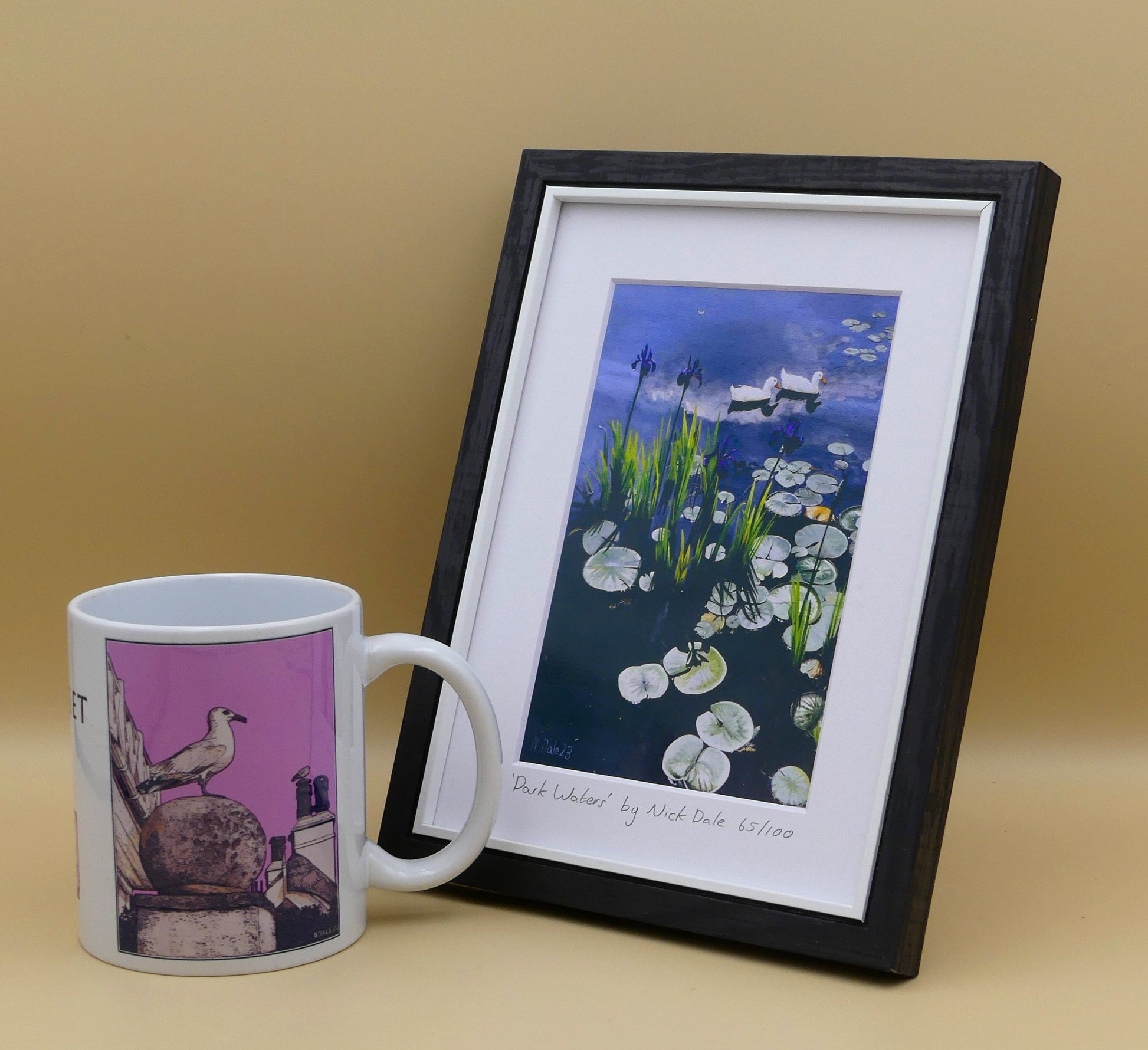Image of a Small Art Print featuring a copy of the painting 'Dark Waters' by Nick Dale. The print depicts lily pads, irises and ducks on a pond at Knightshayes Court, Devon. The print is situated in a black frame next to a mug.