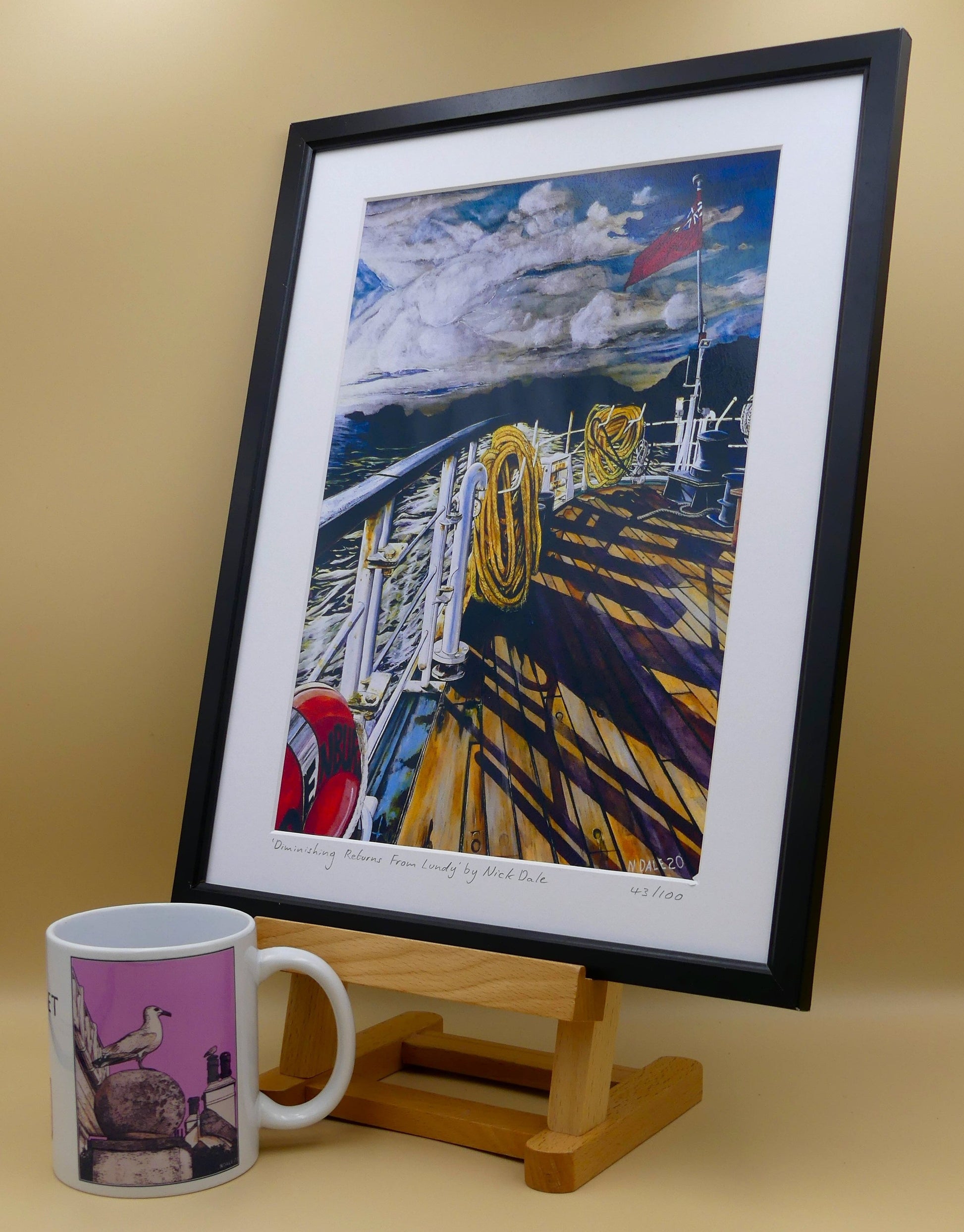 Image of Medium Art Print featuring a copy of the painting 'Diminishing Returns From Lundy' by Nick Dale. The print depicts the stern of the ship MS Oldenburg as it returns to the mainland from Lundy Island, Devon. As the sun goes down it casts long shadows on the deck. whilst the flag flutters in the breeze. The Silhouette of the island can be seen diminishing into the background. The print is situated in a black frame next to a mug.