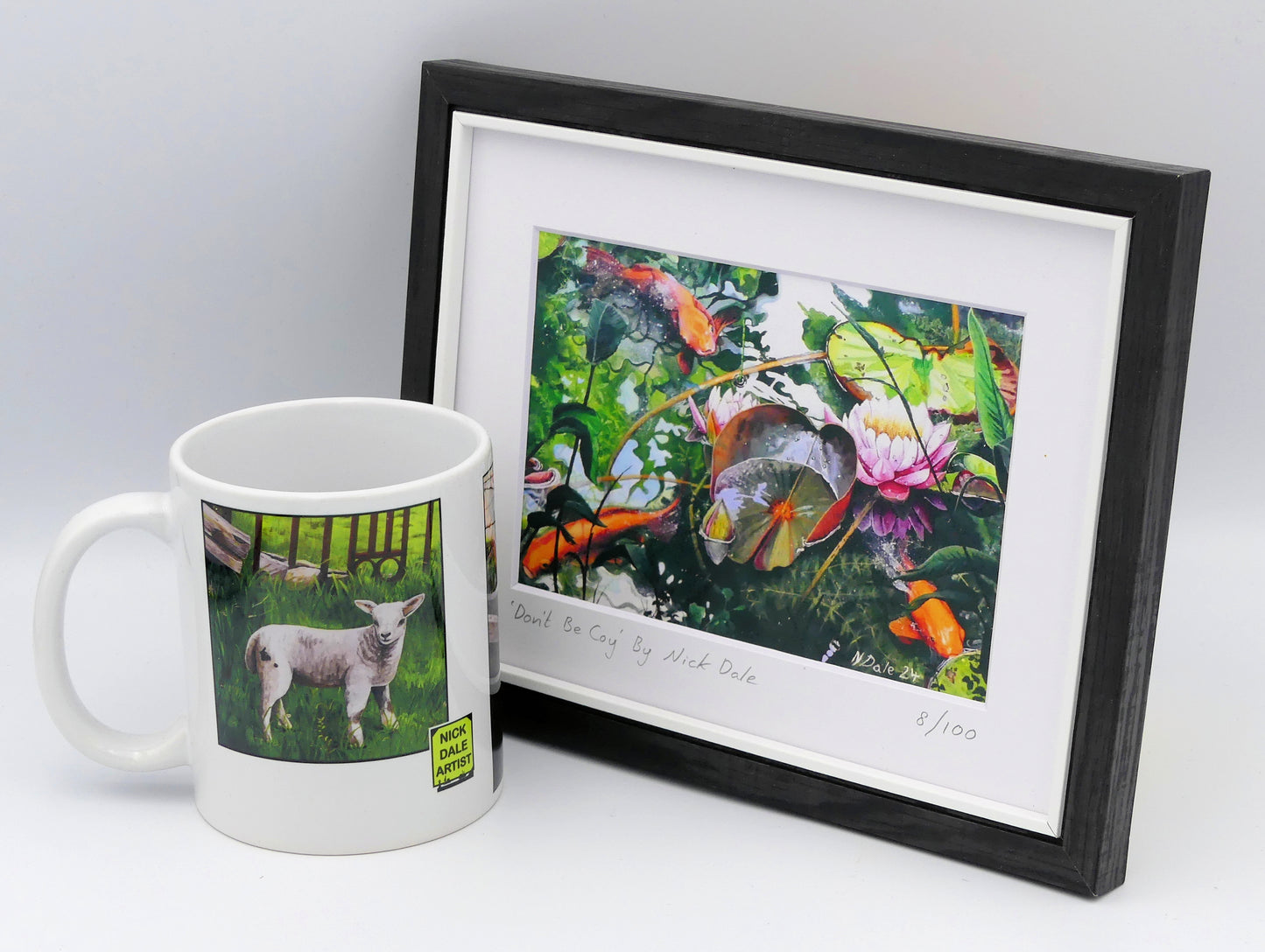 Image of a Small Art Print featuring a copy of the painting 'Don't Be Coy' by Nick Dale. The print depicts 3 koi carp along with water lilies and their flowers. The painting is based on fish and lilies observed at Bennetts Water Gardens in Weymouth, Dorset. The title is a play on the homophones 'Coy' and 'Koi'. The print is situated in a black frame next to a mug.