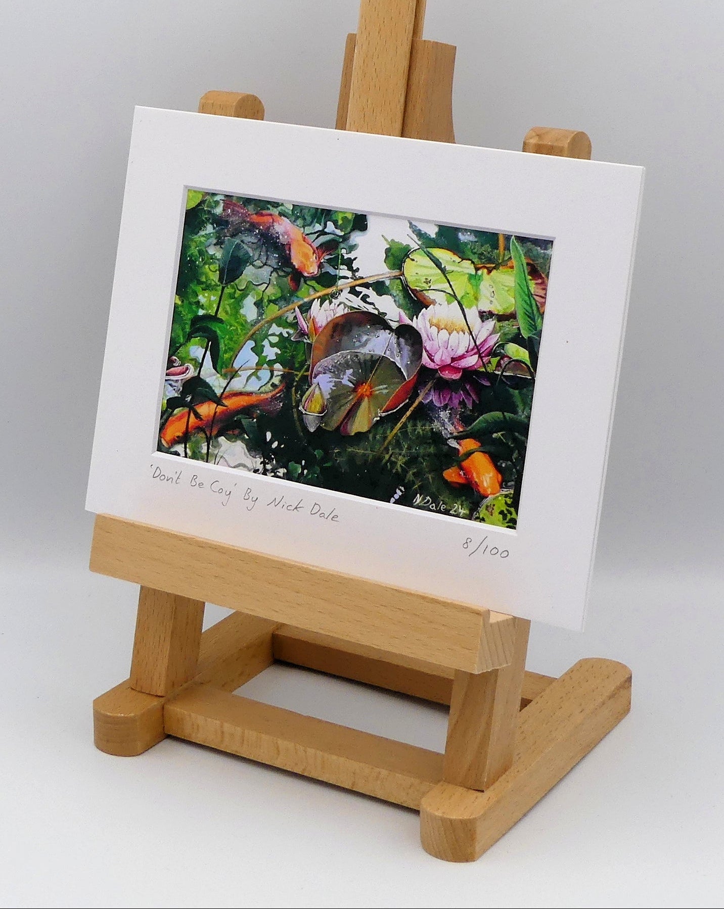 Image of a Small Art Print featuring a copy of the painting 'Don't Be Coy' by Nick Dale. The print depicts 3 koi carp along with water lilies and their flowers. The painting is based on fish and lilies observed at Bennetts Water Gardens in Weymouth, Dorset. The title is a play on the homophones 'Coy' and 'Koi'. The print is situated on a small easel.