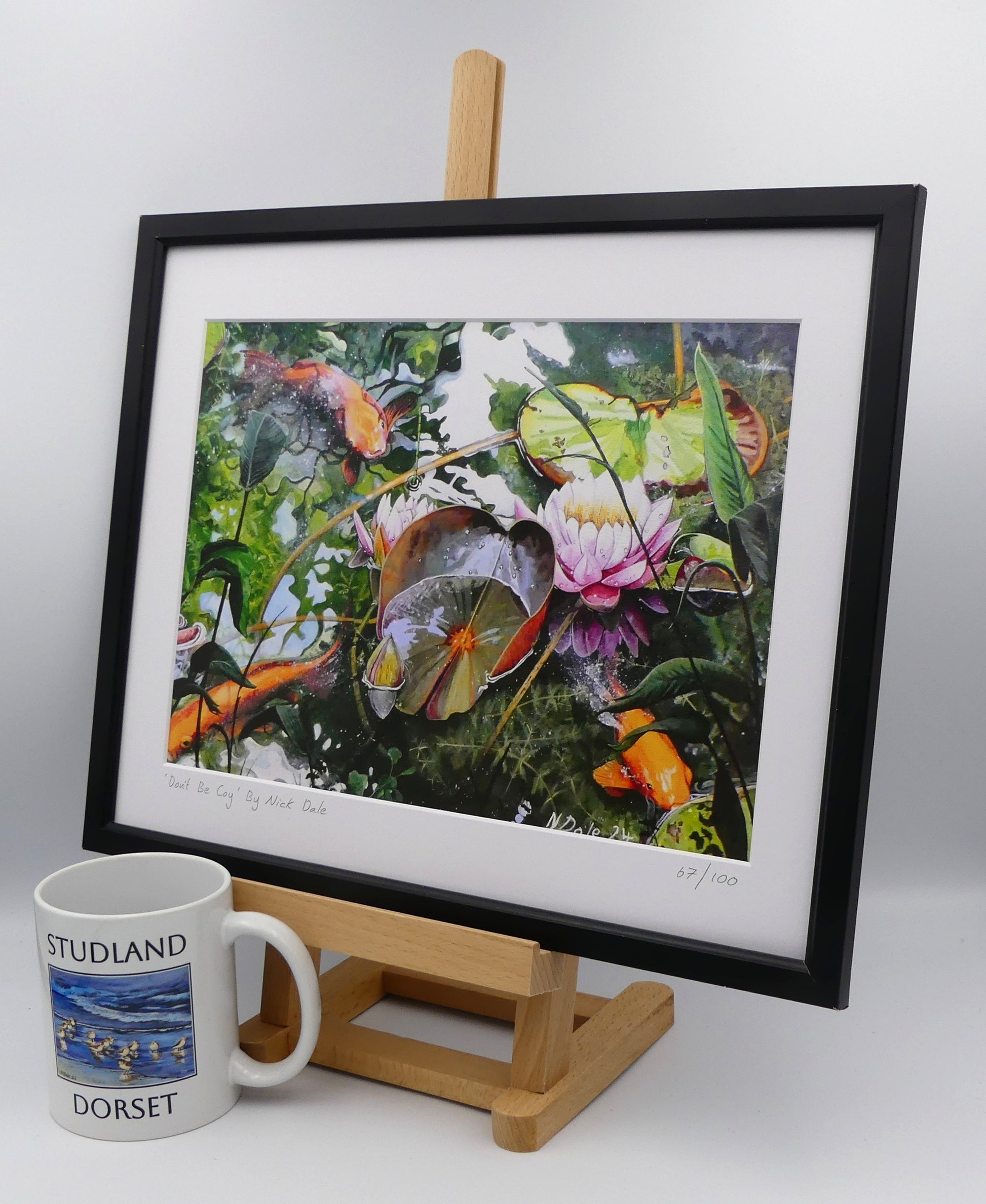 Image of a Medium Art Print featuring a copy of the painting 'Don't Be Coy' by Nick Dale. The print depicts 3 koi carp along with water lilies and their flowers. The painting is based on fish and lilies observed at Bennetts Water Gardens in Weymouth, Dorset. The title is a play on the homophones 'Coy' and 'Koi'. The print is situated on a small easel in a black frame next to a mug.