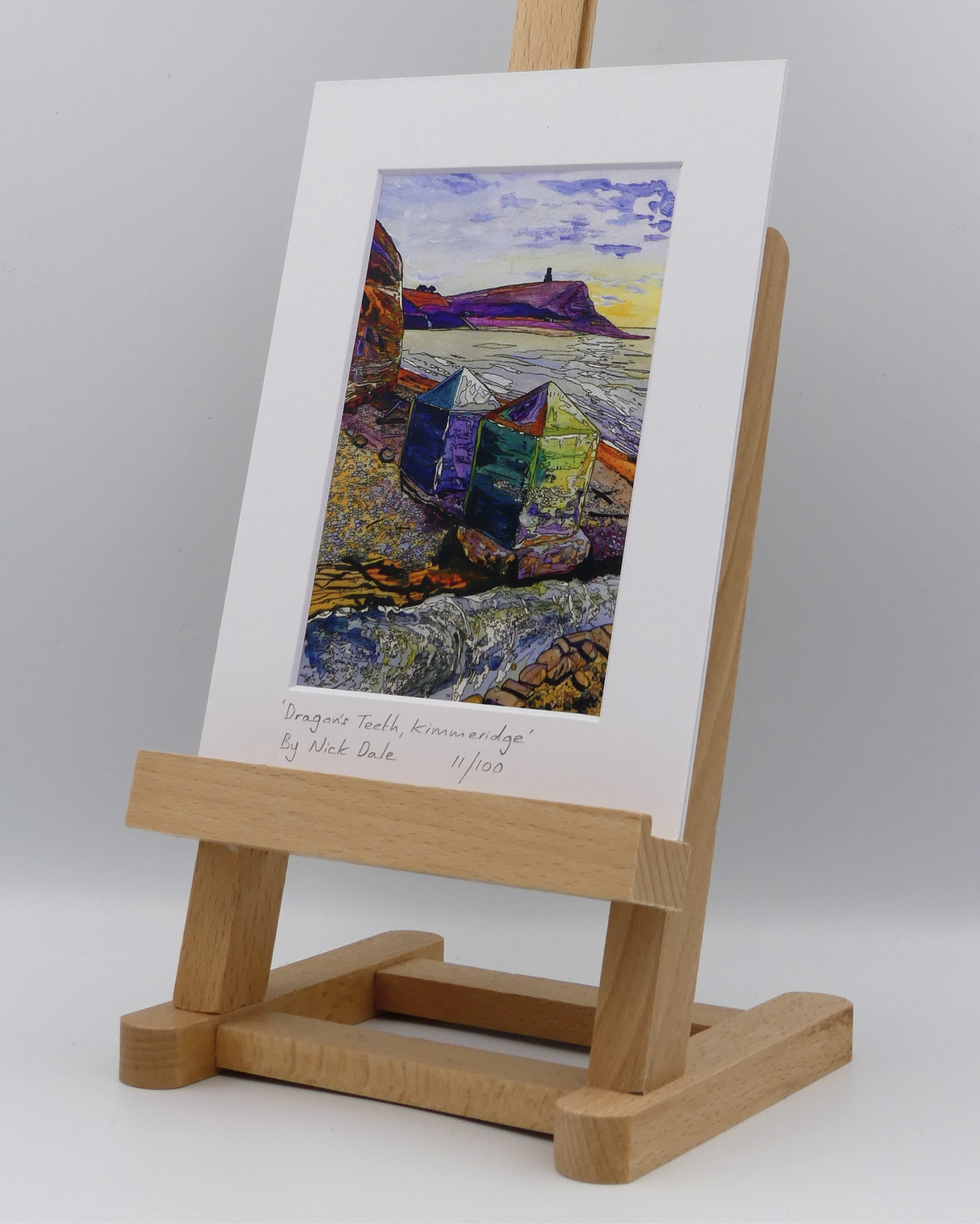 Image of a Small Art Print featuring a copy of the painting 'Dragon's Teeth, Kimmeridge' by Nick Dale. The scene depicts two surviving dragon's teeth anti-tank defences on the beach at Kimmeridge Bay, Dorset. Beyond them can be seen Clavell Tower on the cliff top. The Gaulter Gap forms a relatively easy route off the beach to the cliffs above for any invading force so it was blocked with dragon's teeth and a concrete pillbox during World War II. The print is situated on a small easel.