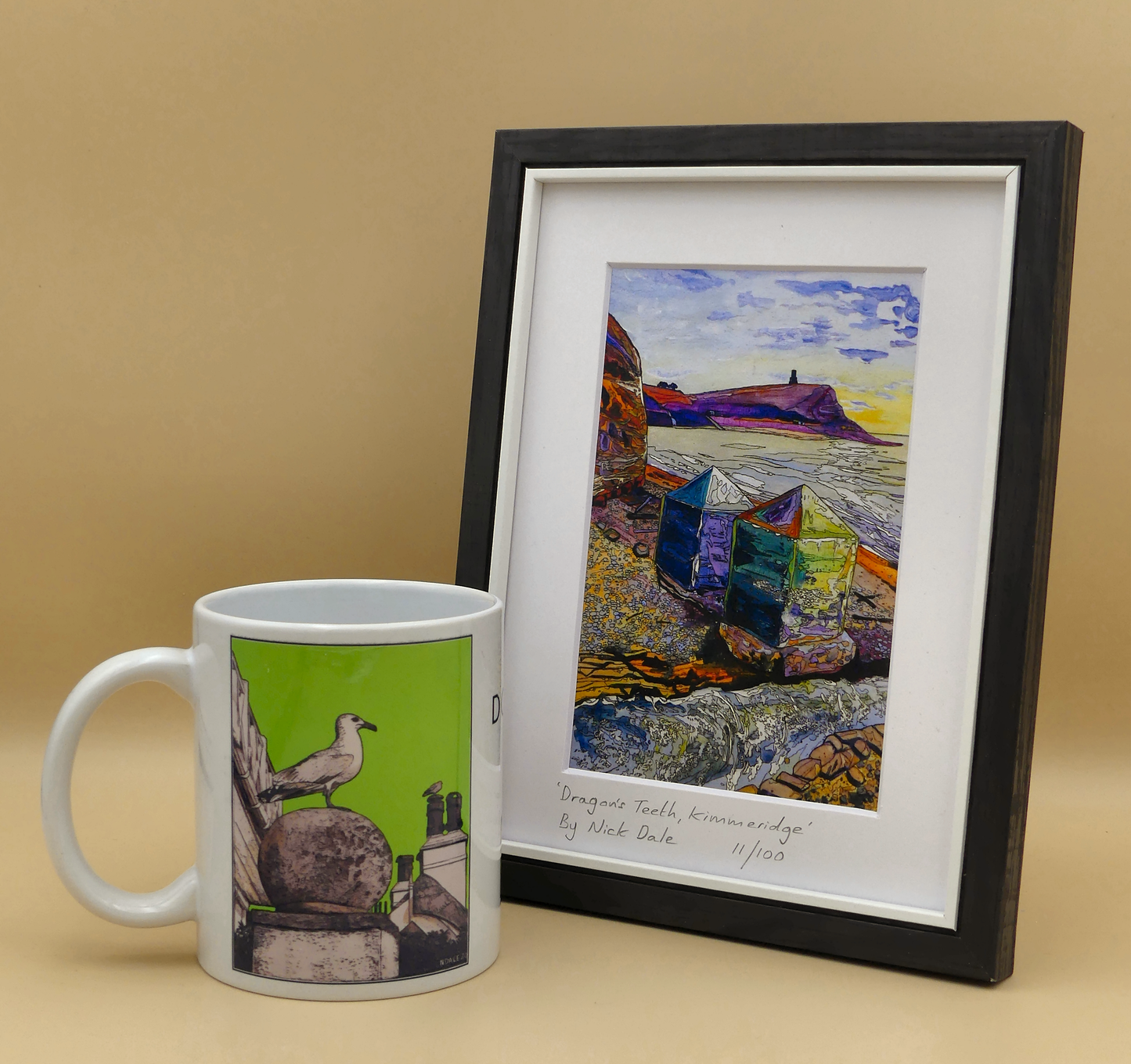 Image of a Small Art Print featuring a copy of the painting 'Dragon's Teeth, Kimmeridge' by Nick Dale. The scene depicts two surviving dragon's teeth anti-tank defences on the beach at Kimmeridge Bay, Dorset. Beyond them can be seen Clavell Tower on the cliff top. The Gaulter Gap forms a relatively easy route off the beach to the cliffs above for any invading force so it was blocked with dragon's teeth and a concrete pillbox during World War II. The print is situated in a black frame next to a mug.