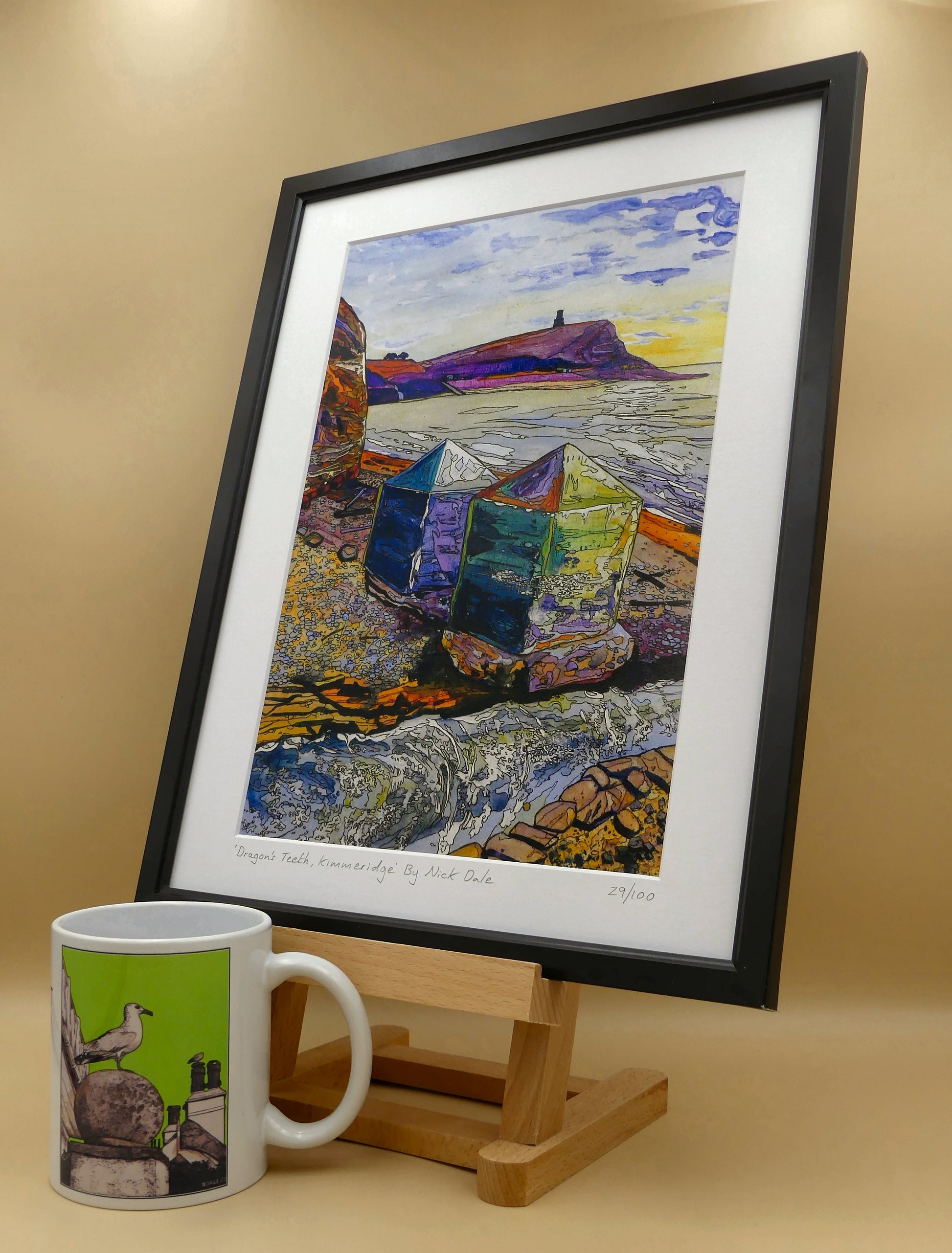 Image of a Medium Art Print featuring a copy of the painting 'Dragon's Teeth, Kimmeridge' by Nick Dale. The scene depicts two surviving dragon's teeth anti-tank defences on the beach at Kimmeridge Bay, Dorset. Beyond them can be seen Clavell Tower on the cliff top. The print is situated on a small easel in a black frame next to a mug.