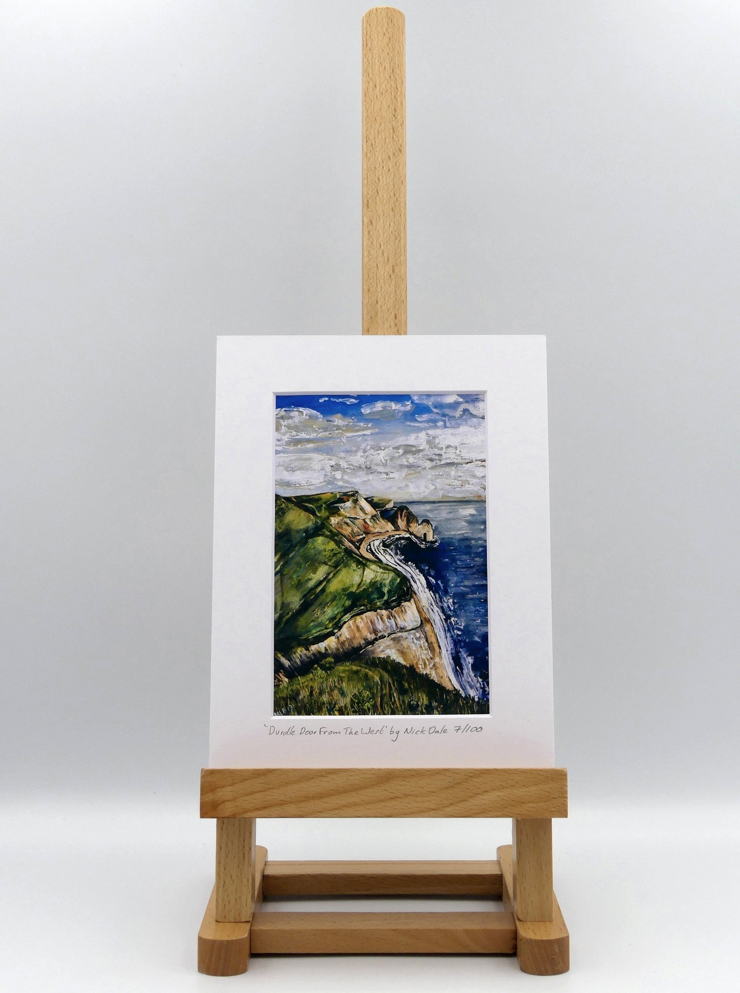 Image of a Small Art Print featuring a copy of the painting 'Durdle Door From The West:' by Nick Dale. The print depicts a view along the Jurassic Coast to the famous Dorset limestone arch landmark, as seen from the coastal path to the west. The print is situated on a small easel.