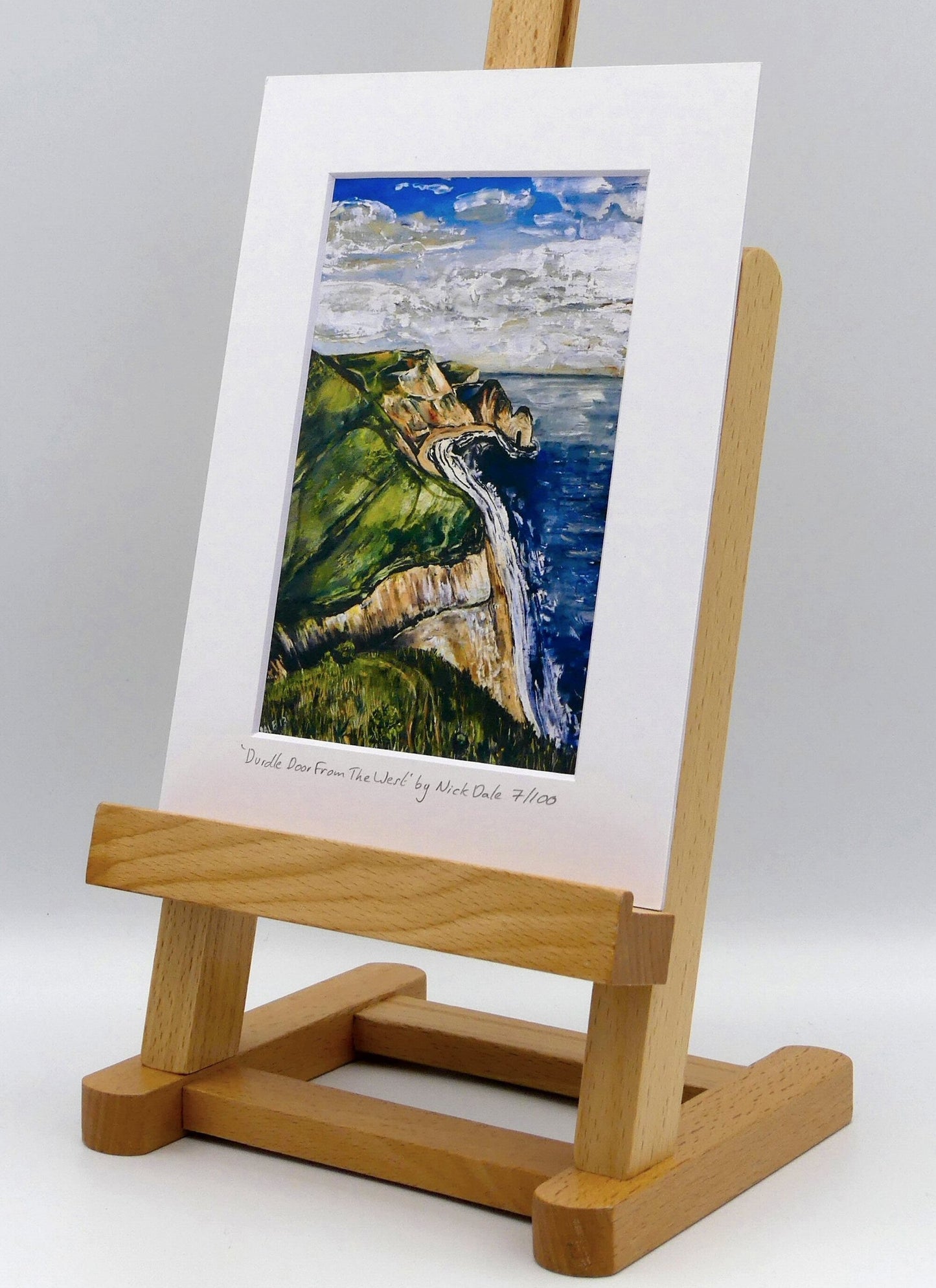Image of a Small Art Print featuring a copy of the painting 'Durdle Door From The West:' by Nick Dale. The print depicts a view along the Jurassic Coast to the famous Dorset limestone arch landmark, as seen from the coastal path to the west. The print is situated on a small easel.
