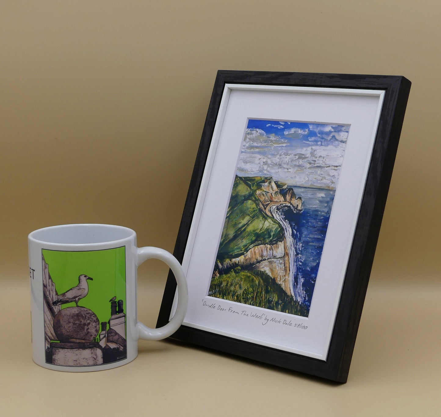 Image of a Small Art Print featuring a copy of the painting 'Durdle Door From The West:' by Nick Dale. The print depicts a view along the Jurassic Coast to the famous Dorset limestone arch landmark, as seen from the coastal path to the west. The print is situated in a black frame next to a mug.