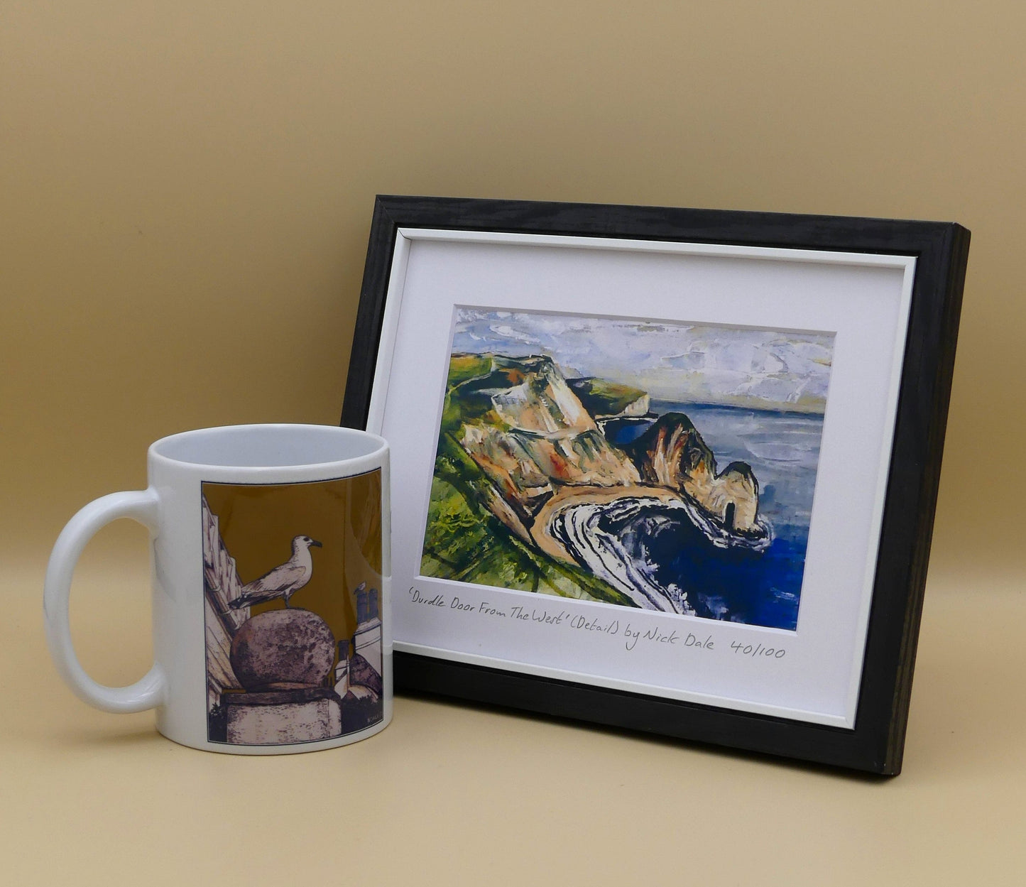 Image of a Small Art Print featuring a copy of a detail from the painting 'Durdle Door From The West:' by Nick Dale. The print depicts a view along the Jurassic Coast to the famous Dorset limestone arch landmark, as seen from the coastal path to the west. The print is situated in a black frame next to a mug.