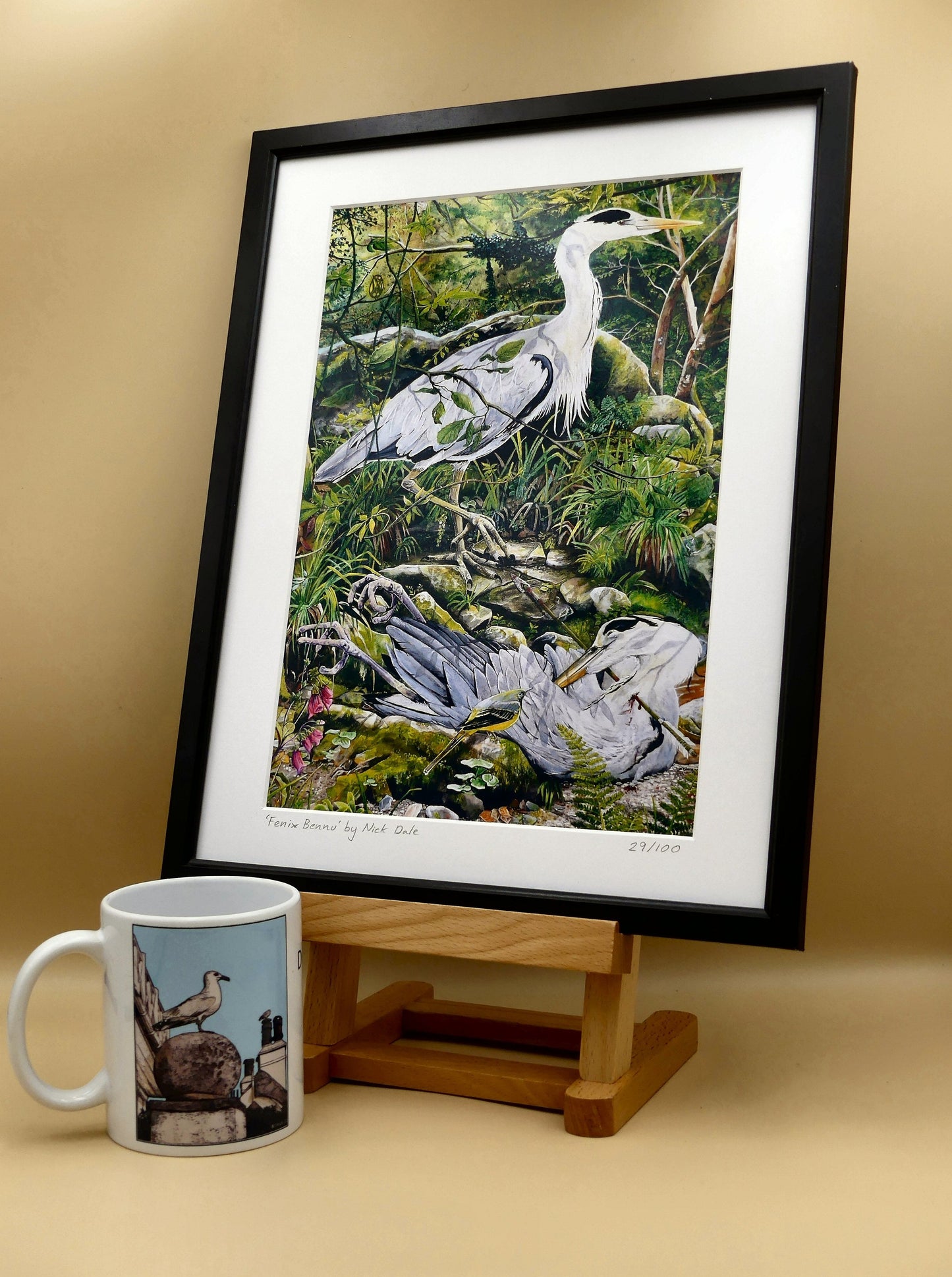 Image of a Medium Art Print featuring a copy of the painting 'Fenix Bennu' by Nick Dale. The print depicts 2 grey herons (1 living & 1 dead) & is named after the Egyptian god Bennu; which may be the origins of the phoenix legend. 'Fenix' is Anglo-Saxon for phoenix & the scene has been transposed to a river in Devon. The blood of the dead heron is smoking where it drips on the ground; suggesting that this is indeed a phoenix. The print is situated in a black frame next to a mug.