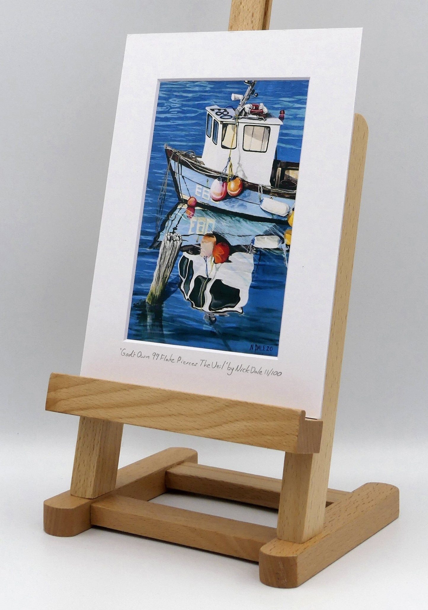 Image of a Small Art Print featuring a copy of the painting 'God's Own 99 Flake Pierces The Veil' by Nick Dale. The print depicts a fishing boat at rest within the harbour of Lyme Regis, Dorset. The title is based on the similarity of the wooden pile to a chocolate flake as inserted into an ice cream cone. This seemed appropriate given the draw of the town to tourists. The print is situated on a small easel.