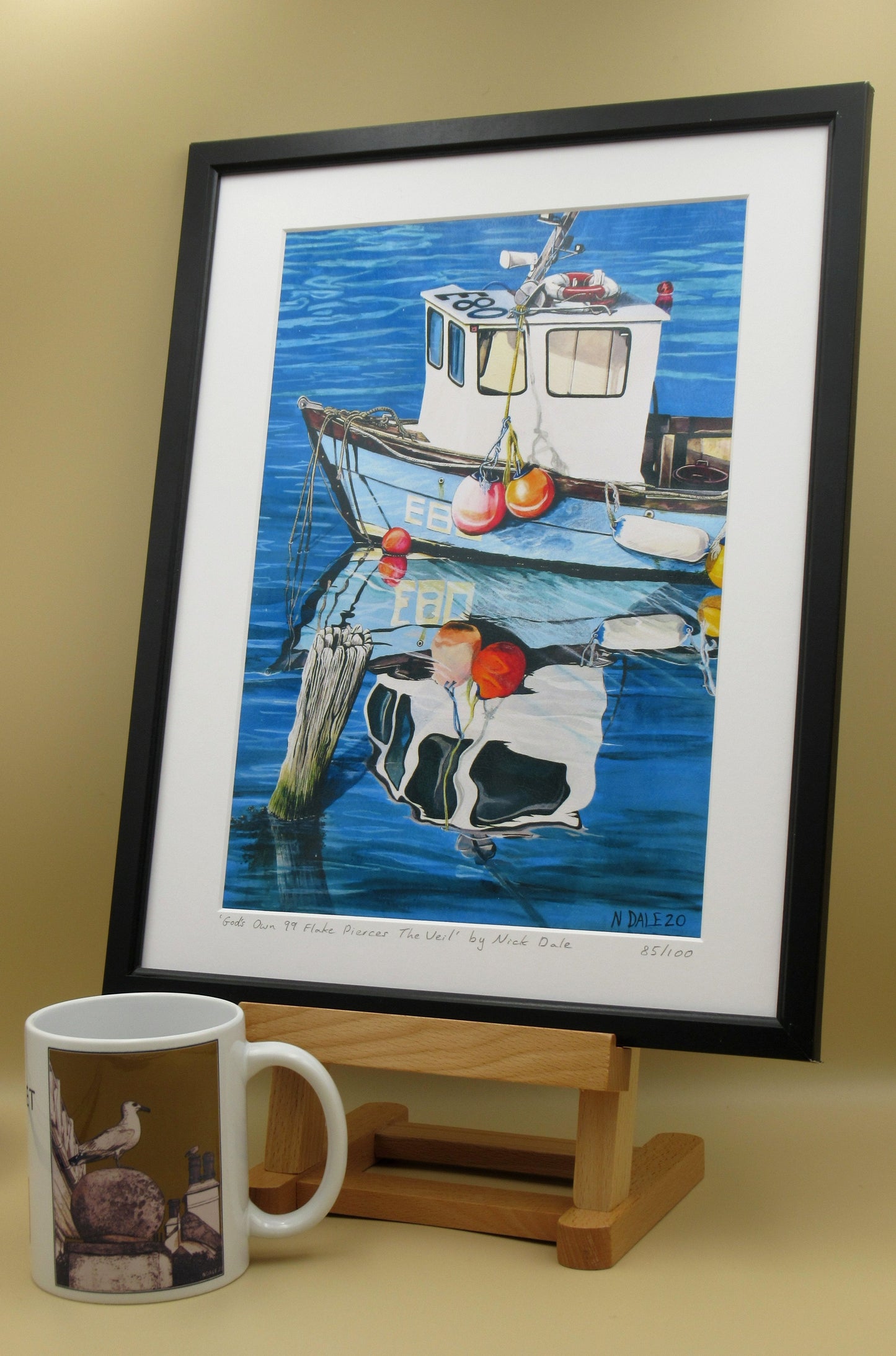 Image of a Medium Art Print featuring a copy of the painting 'God's Own 99 Flake Pierces The Veil' by Nick Dale. The print depicts a fishing boat at rest within the harbour of Lyme Regis, Dorset. The title is based on the similarity of the wooden pile to a chocolate flake as inserted into an ice cream cone. This seemed appropriate given the draw of the town to tourists. The print is situated in a black frame next to a mug.
