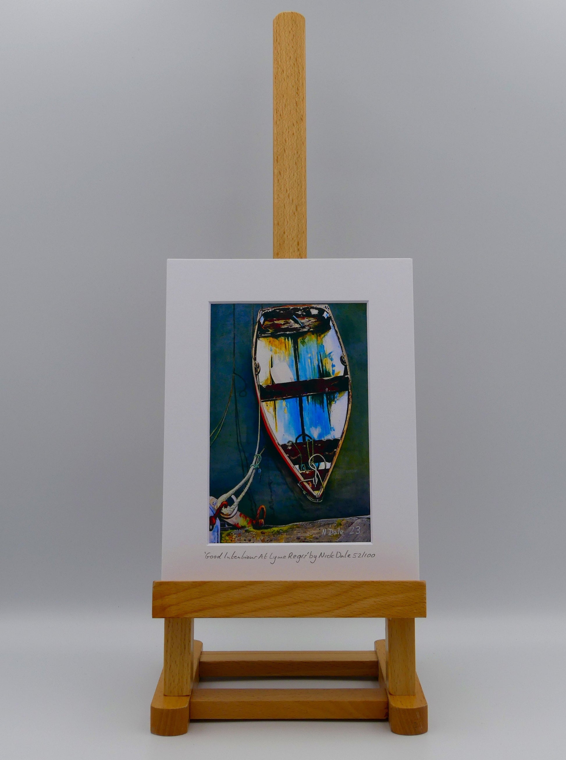 Image of a Small Art Print featuring a copy of the painting 'Good Intentions at Lyme Regis' by Nick Dale. The print depicts a small wooden rowing boat tied up in Lyme Regis harbour, Dorset. The print is situated on a small easel.