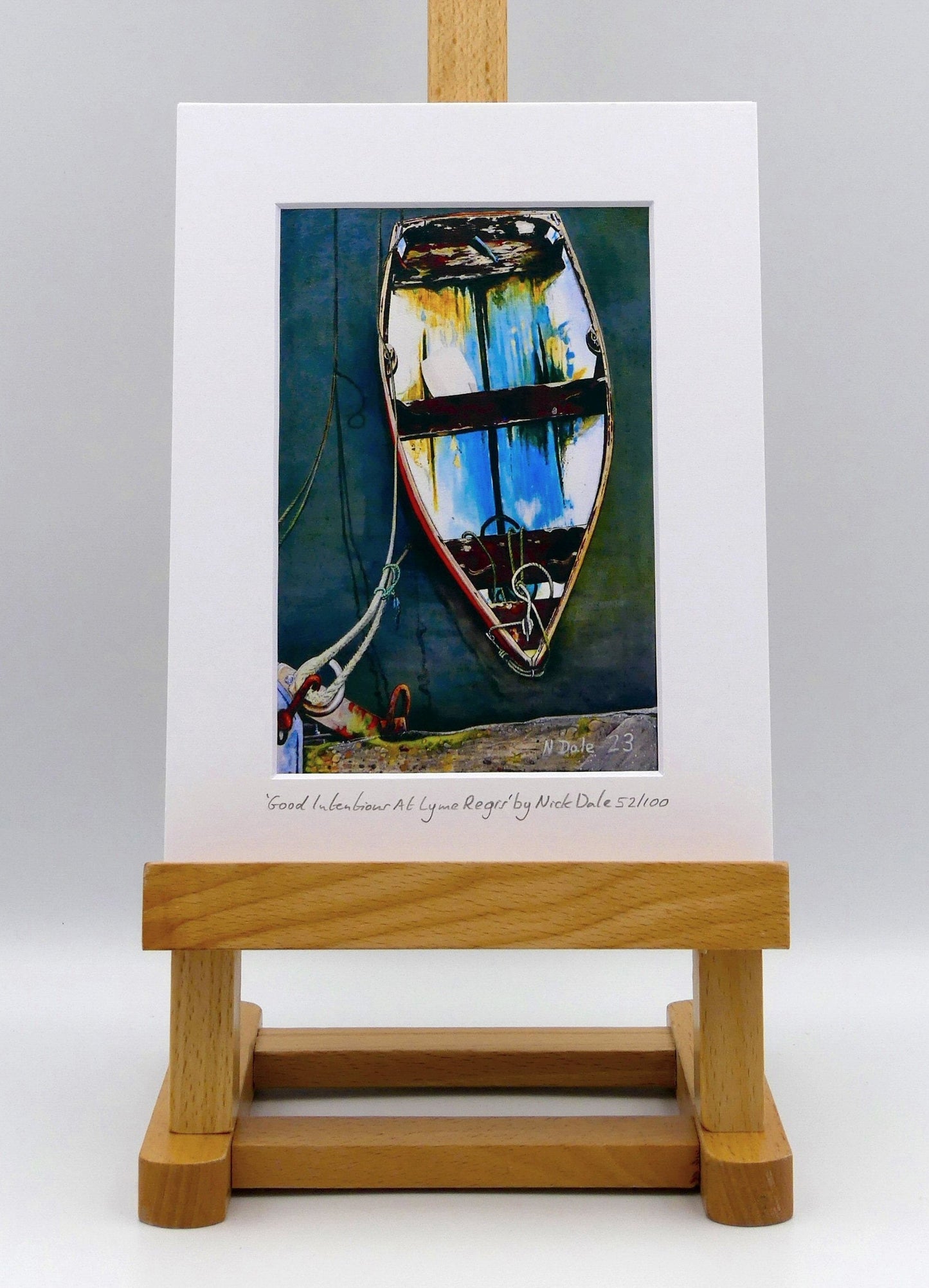 Image of a Small Art Print featuring a copy of the painting 'Good Intentions at Lyme Regis' by Nick Dale. The print depicts a small wooden rowing boat tied up in Lyme Regis harbour, Dorset. The print is situated on a small easel.