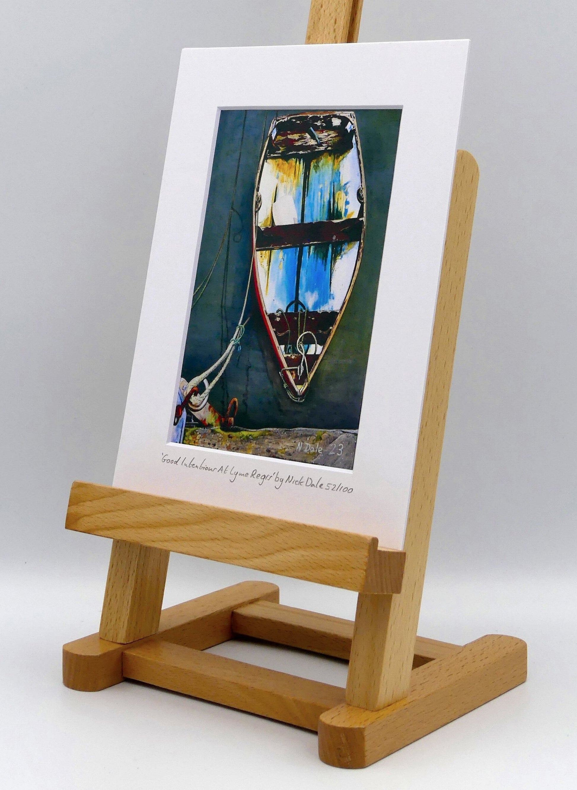 Image of a Small Art Print featuring a copy of the painting 'Good Intentions at Lyme Regis' by Nick Dale. The print depicts a small wooden rowing boat tied up in Lyme Regis harbour, Dorset. The print is situated on a small easel.