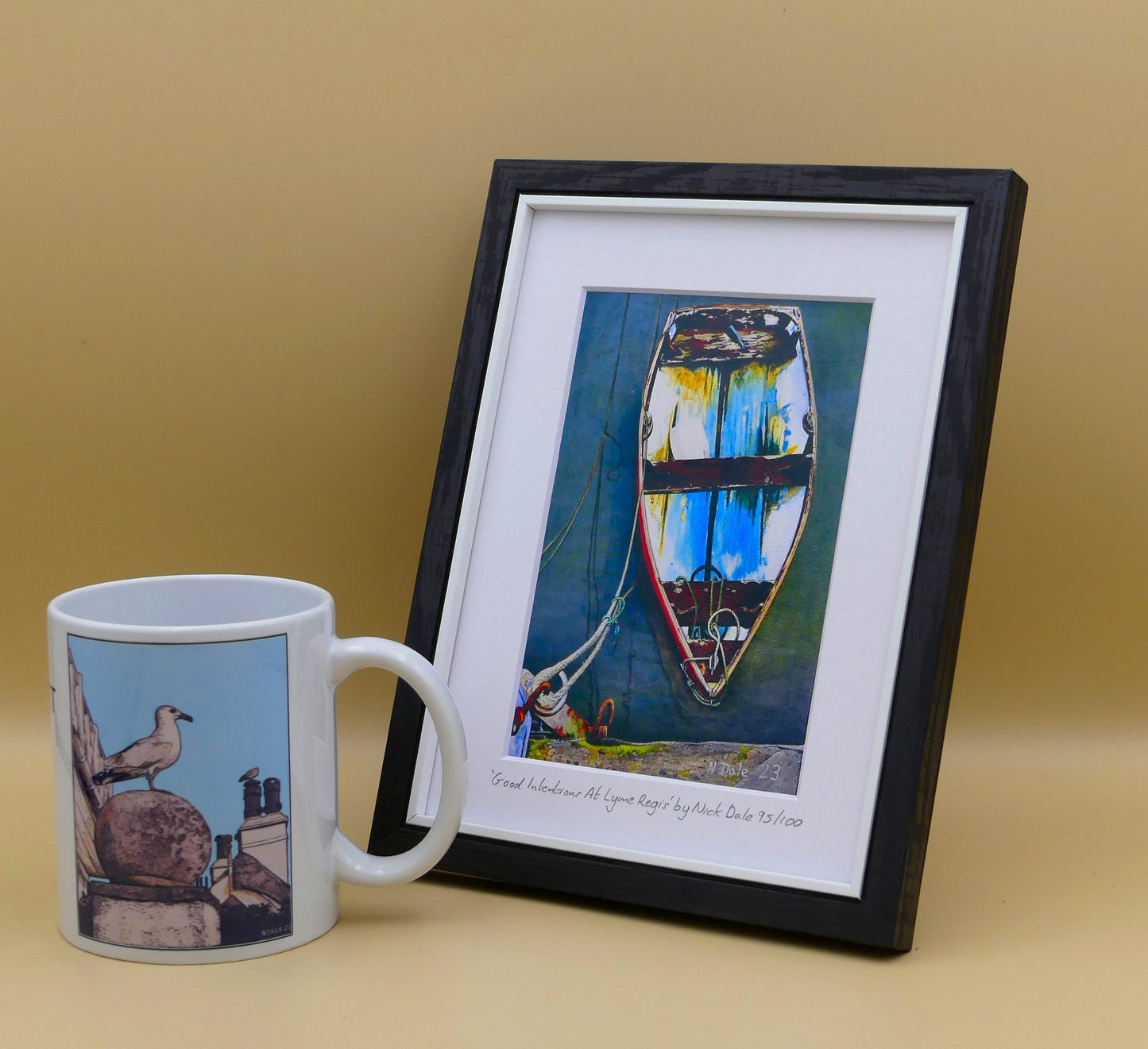 Image of a Small Art Print featuring a copy of the painting 'Good Intentions at Lyme Regis' by Nick Dale. The print depicts a small wooden rowing boat tied up in Lyme Regis harbour, Dorset. The print is situated in a black frame next to a mug.