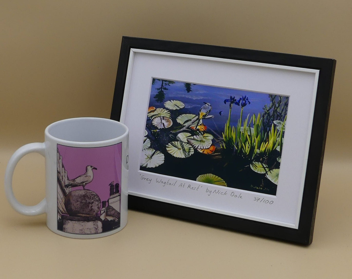 Image of Small Art Print featuring a copy of the painting 'Grey Wagtail At Rest' by Nick Dale. The print depicts a grey wagtail bird at rest, perched on top of a submerged stick above a pond. Whilst the grey wagtail was observed at Athelhampton House in Dorset the pond's vegetation is more closely based on a pond at Knightshayes Court in Devon. The print is situated in a black frame next to a mug.
