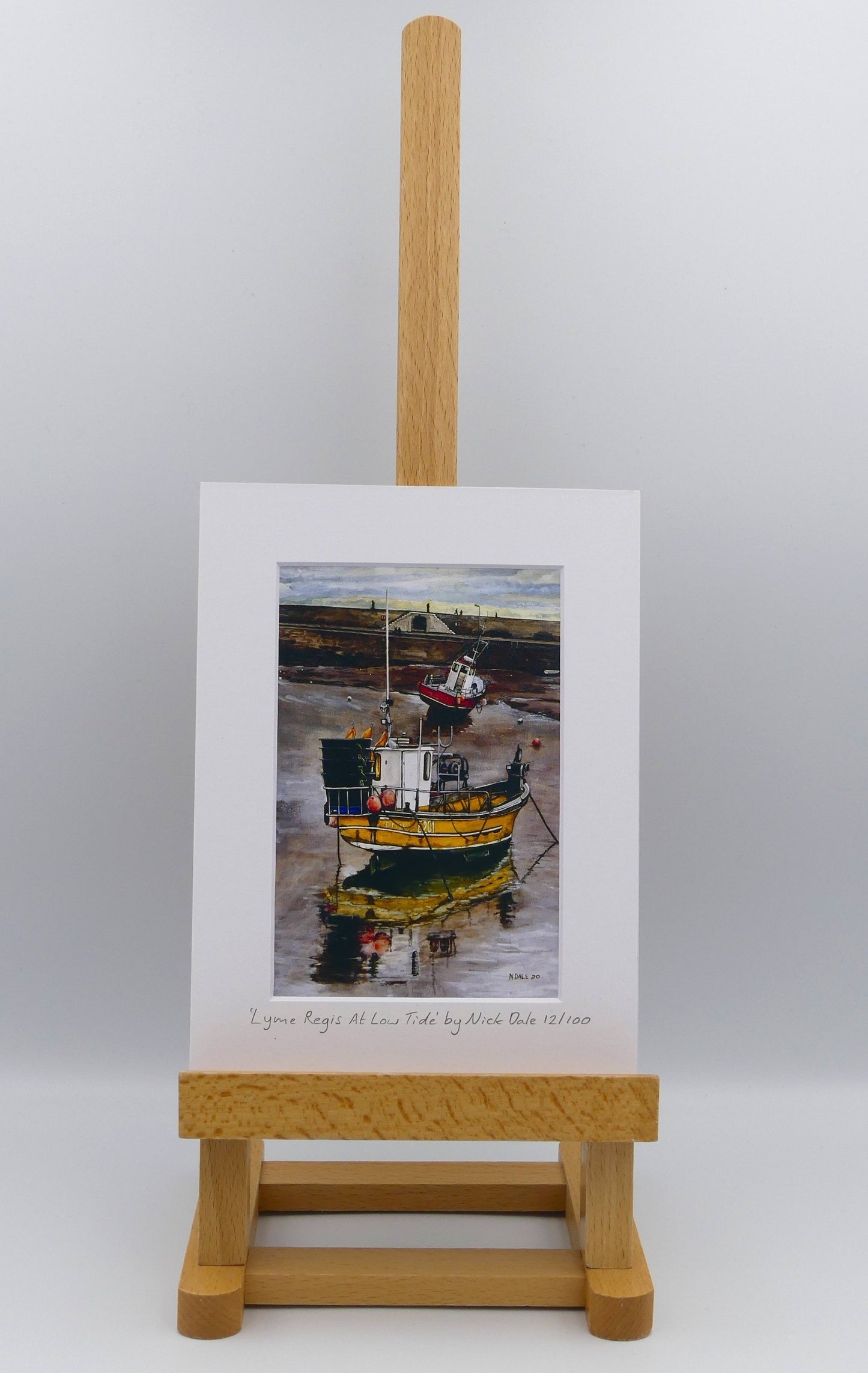 Image of Small Art Print featuring a copy of the painting 'Lyme Regis at Low Tide' by Nick Dale. The print depicts a sunset where the fishing vessels rest on the floor of Lyme Regis harbour whilst the tourists enjoy the view from the Cobb. The print is situated on a small easel.