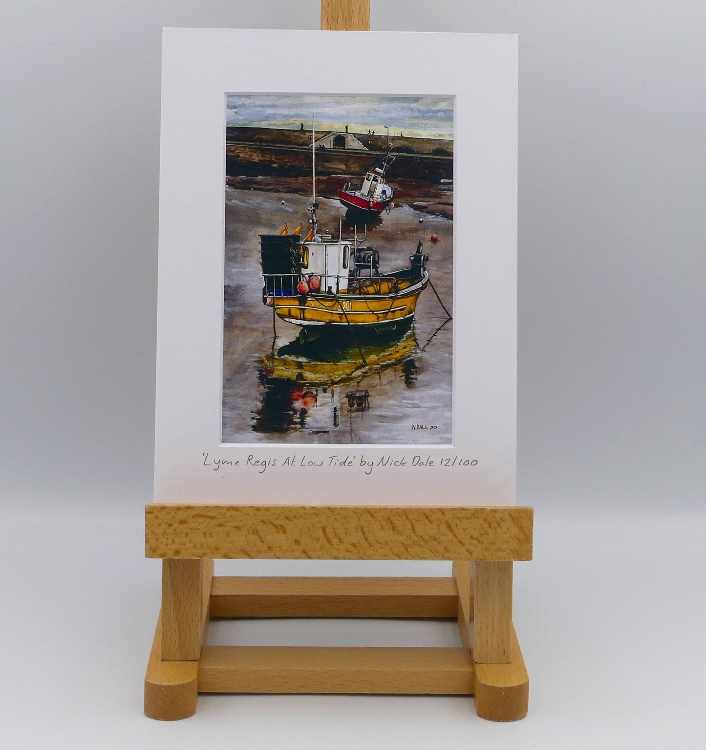 Image of Small Art Print featuring a copy of the painting 'Lyme Regis at Low Tide' by Nick Dale. The print depicts a sunset where the fishing vessels rest on the floor of Lyme Regis harbour whilst the tourists enjoy the view from the Cobb. The print is situated on a small easel.