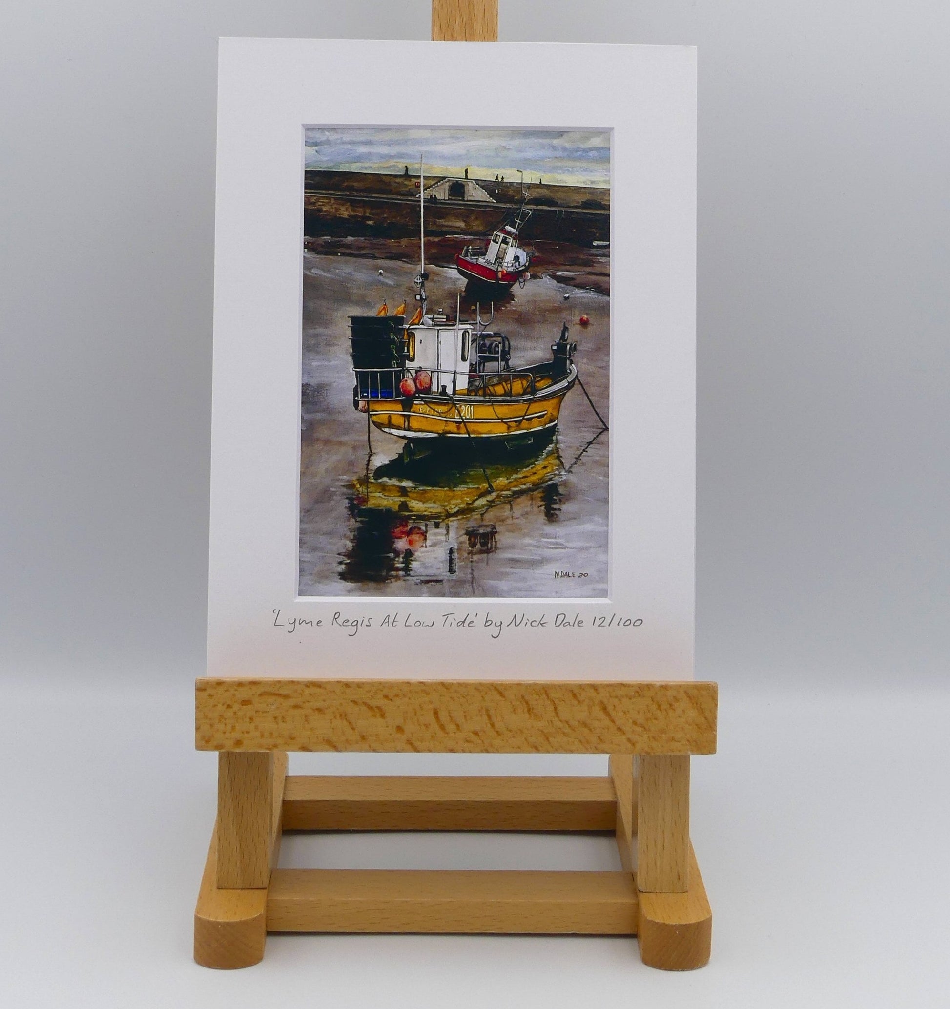 Image of Small Art Print featuring a copy of the painting 'Lyme Regis at Low Tide' by Nick Dale. The print depicts a sunset where the fishing vessels rest on the floor of Lyme Regis harbour whilst the tourists enjoy the view from the Cobb. The print is situated on a small easel.