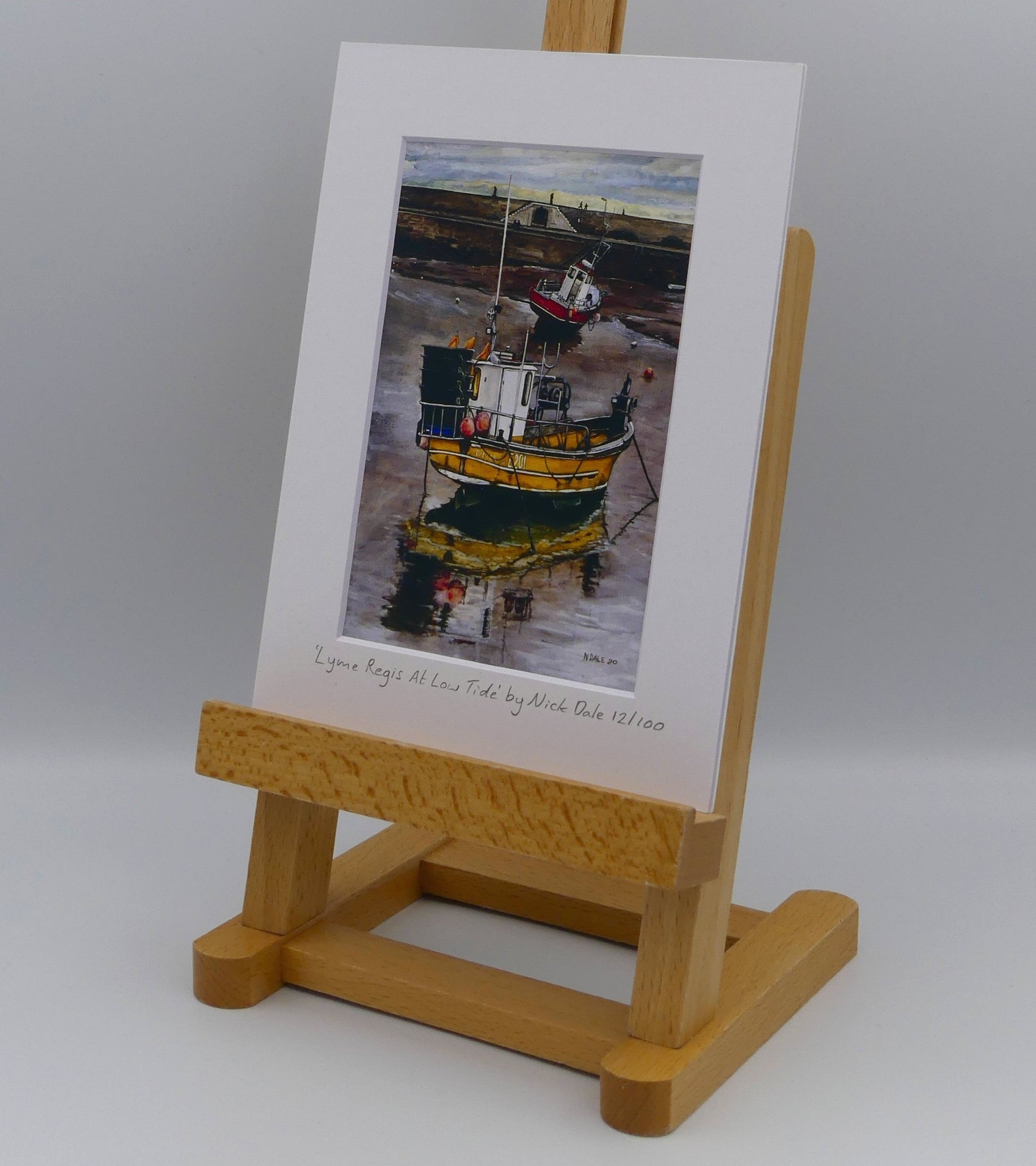 Image of Small Art Print featuring a copy of the painting 'Lyme Regis at Low Tide' by Nick Dale. The print depicts a sunset where the fishing vessels rest on the floor of Lyme Regis harbour whilst the tourists enjoy the view from the Cobb. The print is situated on a small easel.