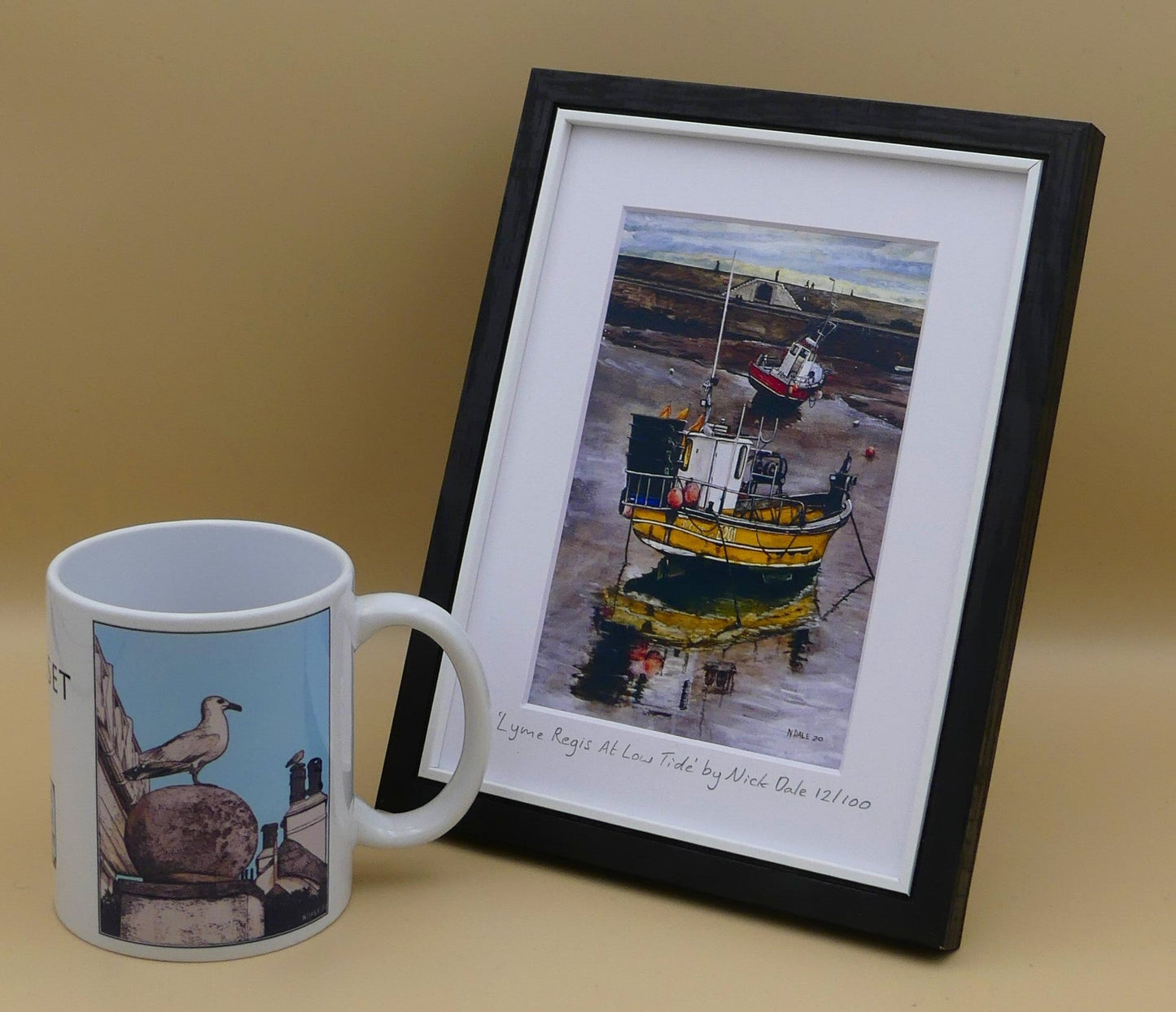 mage of Small Art Print featuring a copy of the painting 'Lyme Regis at Low Tide' by Nick Dale. The print depicts a sunset where the fishing vessels rest on the floor of Lyme Regis harbour whilst the tourists enjoy the view from the Cobb. The print is situated in a black frame next to a mug.