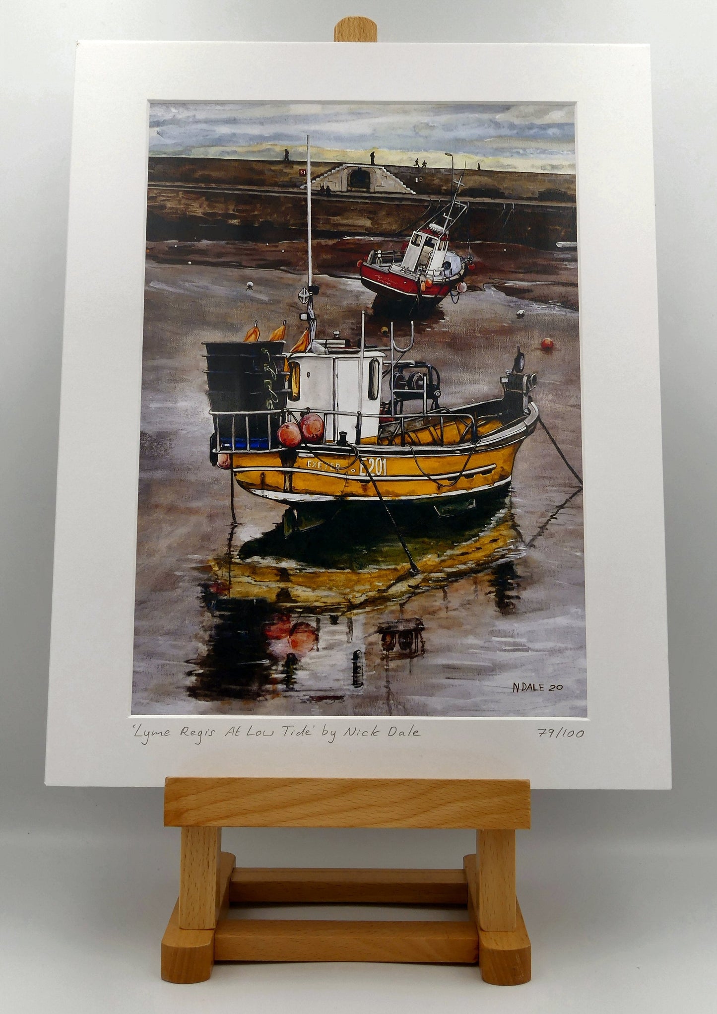 Image of Medium Art Print featuring a copy of the painting 'Lyme Regis at Low Tide' by Nick Dale. The print depicts a sunset where the fishing vessels rest on the floor of Lyme Regis harbour whilst the tourists enjoy the view from the Cobb. The print is situated on a small easel.